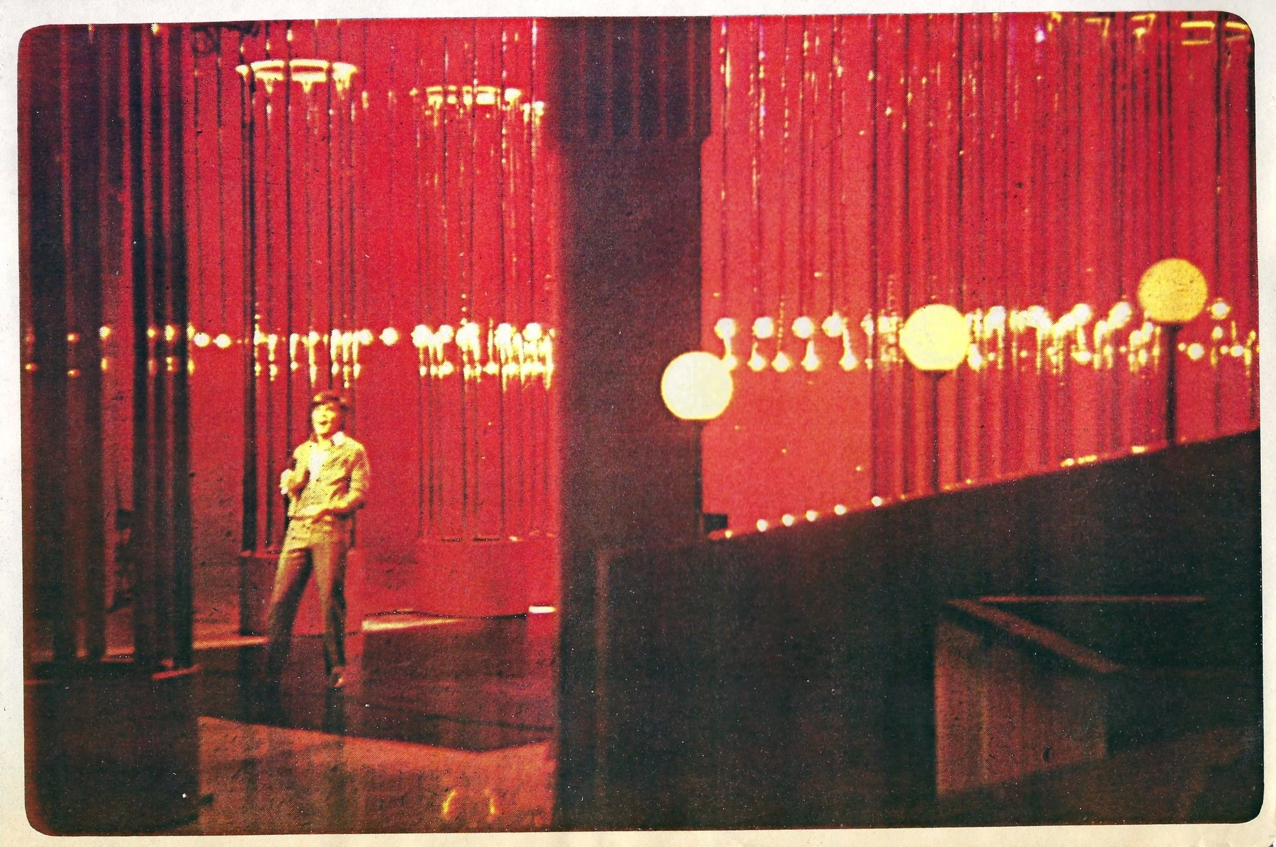 A set photograph from the John Davidson Show, standing with arms crossed in front of a large, illuminated red curtain with circular light fixtures.