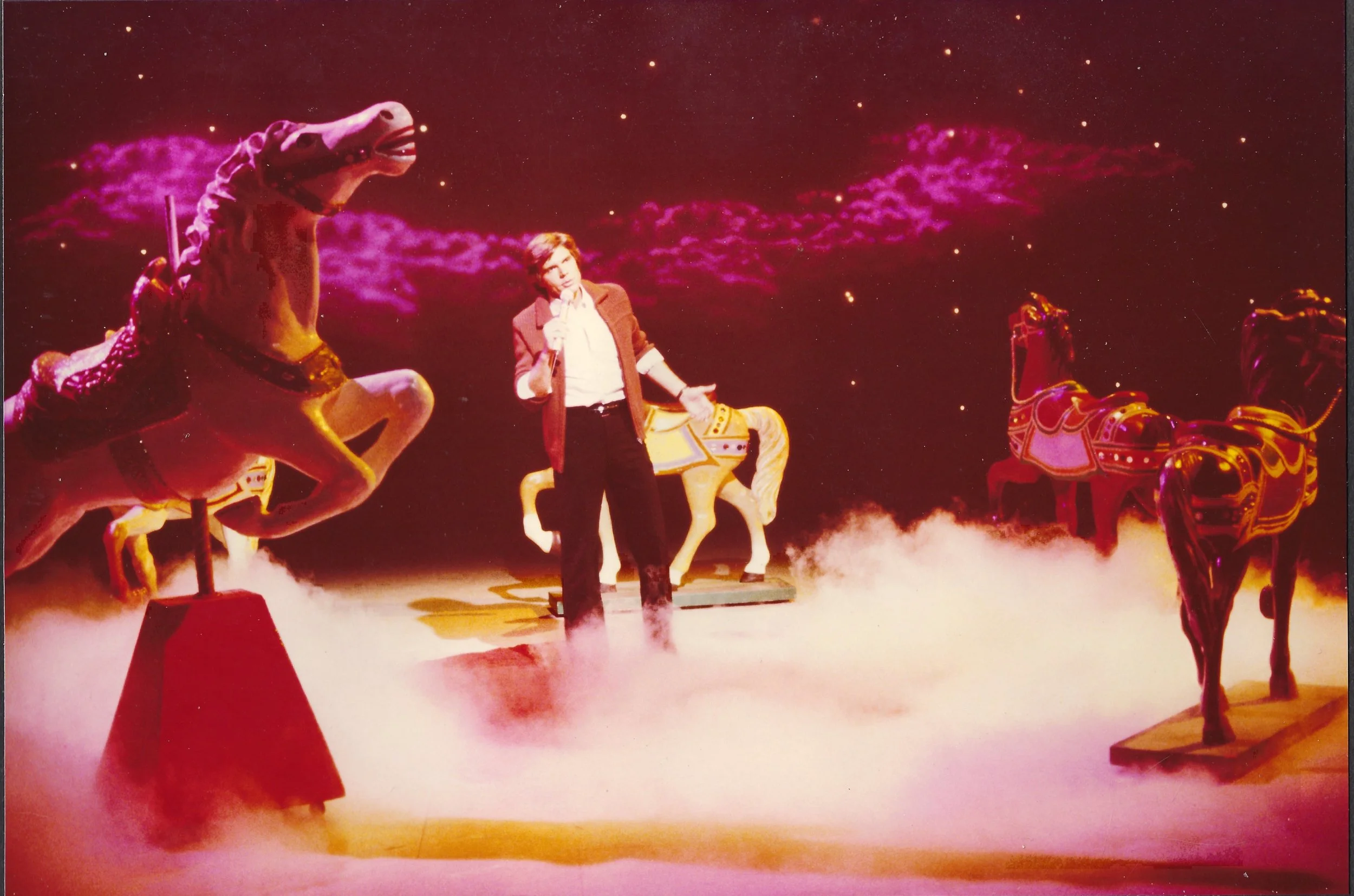 A set photograph from the John Davidson Show, in a brown blazer and white shirt holding a microphone, standing on stage with carousel horses and purple clouds in the background.