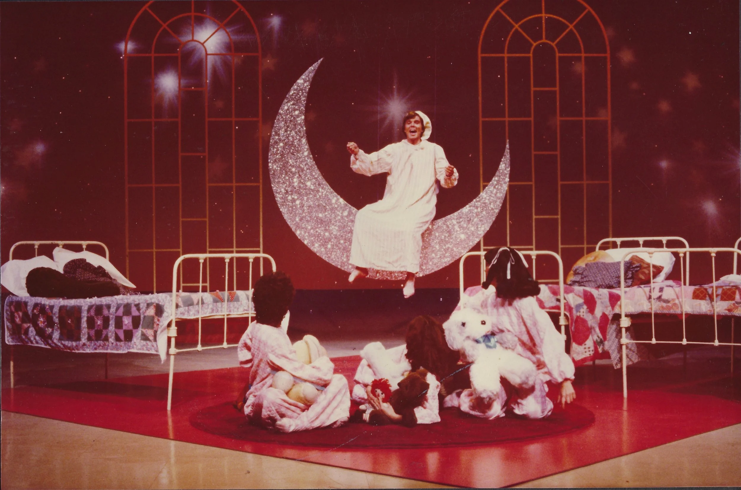 A theatrical scene with a person dressed in pajamas sitting on a large crescent moon prop. Several children and plush toys are gathered around, watching the performance. The background features a starry night sky with clouds and two tall window-like structures. The beds and bedding on stage suggest a fantasy or dream setting.