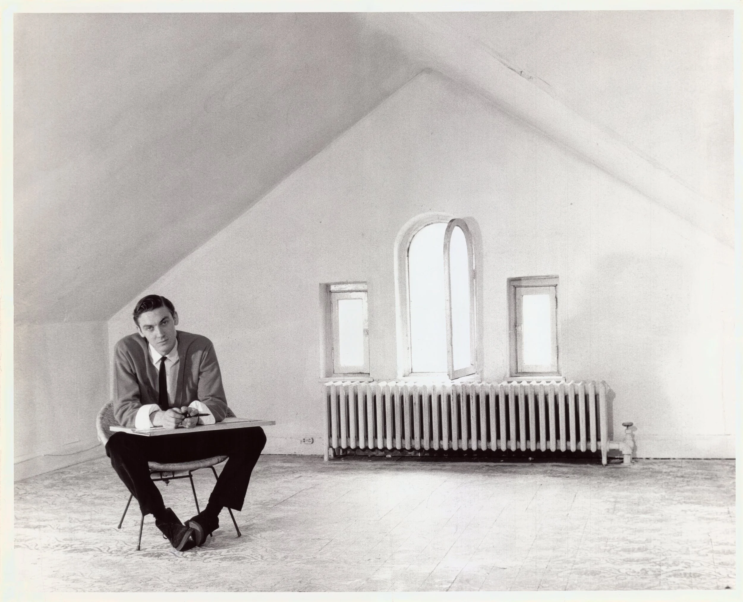 A black-and-white photo of a man sitting on a chair in a spacious, well-lit room with high ceilings, arched windows, and a radiator. The man is holding a cigarette and wearing a suit, with a serious expression on his face.