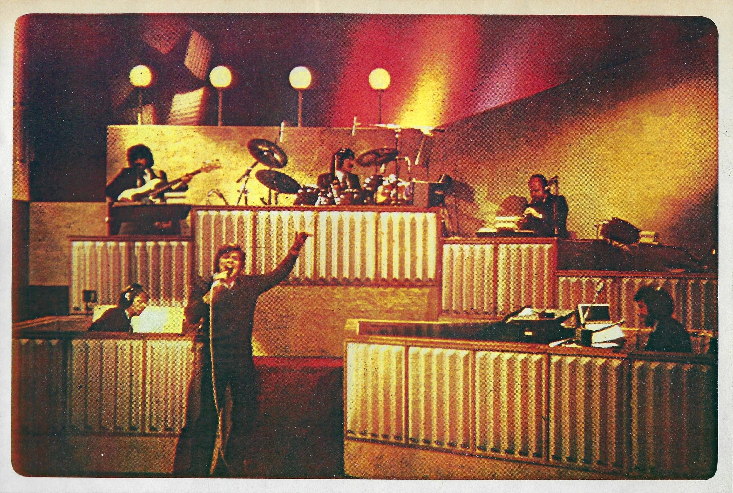 An indoor concert scene with a band performing on stage. The band includes a guitarist, a drummer, and two keyboard players. John Davidson is standing in front of the stage, raising his arm to the audience.
