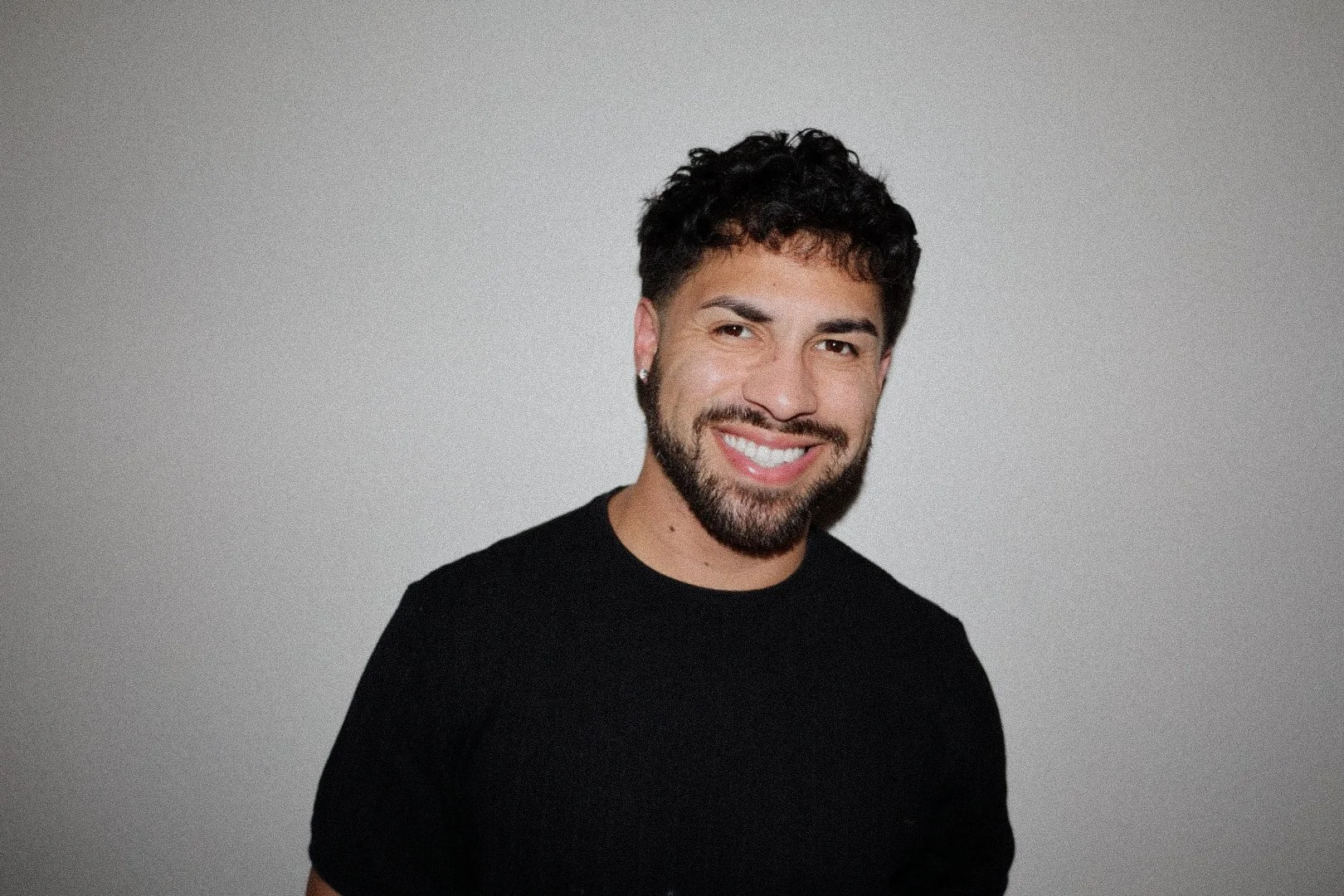 Portrait of a smiling man with dark curly hair, beard, and mustache wearing a black shirt against a plain light gray background.