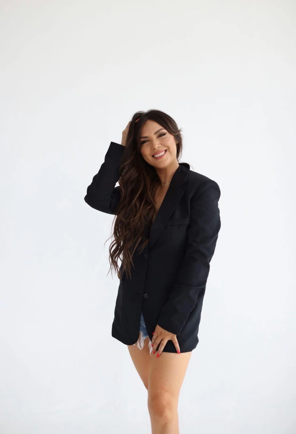 A young woman with long, wavy dark hair, wearing a black blazer and denim shorts with lace trim, smiling and touching her hair on a plain white background.