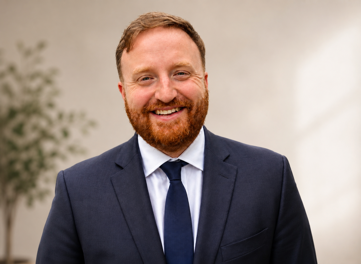 Smiling man in a dark suit, white shirt, and dark tie, with red hair and beard, standing indoors with a blurred plant in the background.