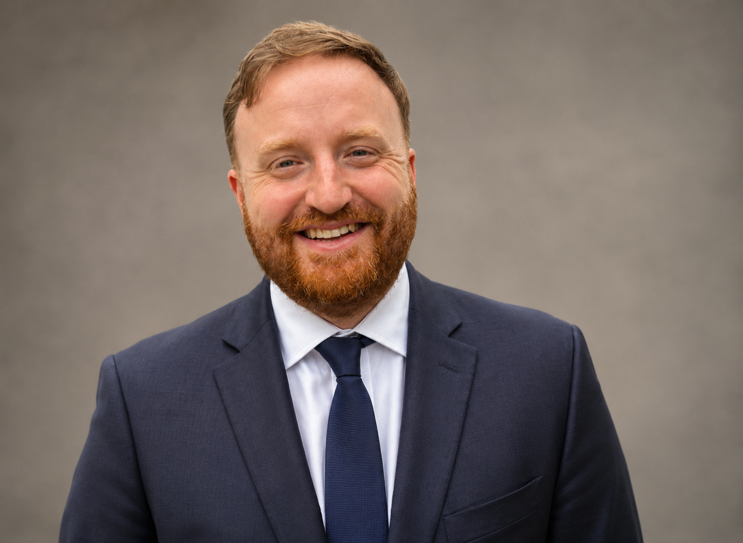 A smiling man with red hair and beard in a navy suit, white shirt, and navy tie, standing against a plain gray background.