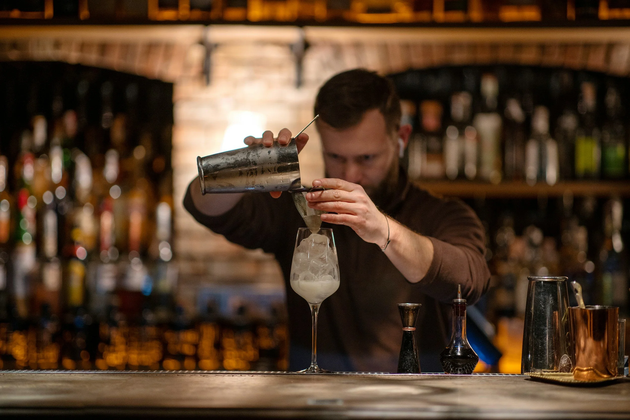 Bartender pouring a drink into a glass with ice at a bar, with bar bottles and a brick wall in the background.