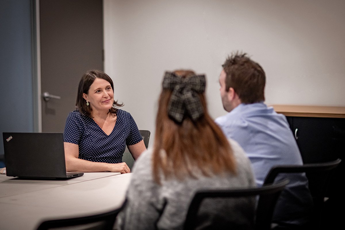 Three people in a meeting, a woman on the left facing the camera and two people on the right with their backs to the camera, a woman with a bow in her hair and a man with dark hair, in a conference room with a laptop on the table.