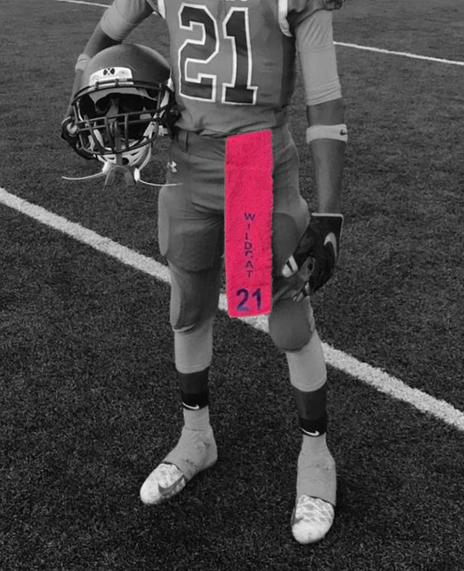 A young football player in a uniform, holding a helmet, standing on a sports field with a pink towel labeled 'Wildcat 21' around their waist.