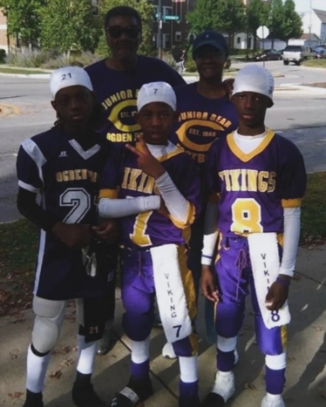 A group of five young boys and one adult man posing for a photo outdoors. The boys and the man are wearing sports uniforms and baseball caps; each has a different number on their cap and jersey. The boys' jerseys say 'VIKINGS' and the adult's shirt says 'JUNIOR BAND.' They seem to be part of a sports team, possibly football or baseball, standing on a sidewalk with trees and houses in the background.