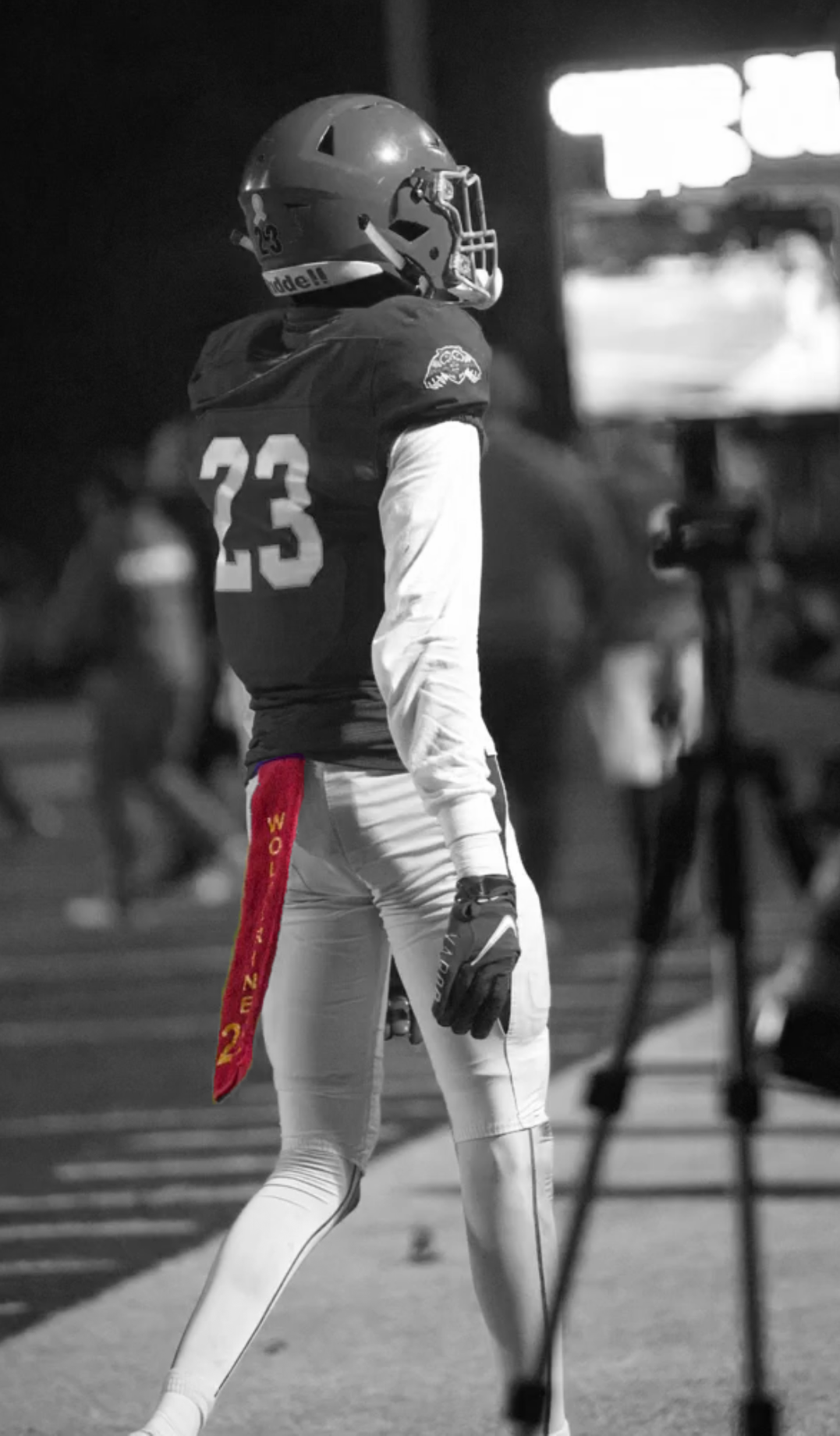A football player wearing a helmet, football uniform with number 23, standing on the sideline during a game at night with a custom colorful towel.