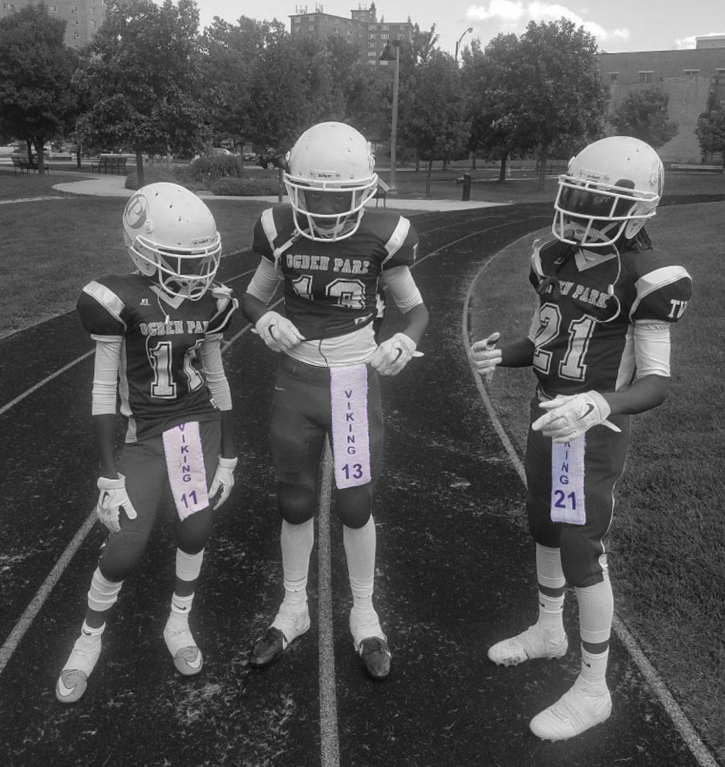 Three young football players in uniform standing on a track field, wearing helmets and pads, preparing for a game or practice.