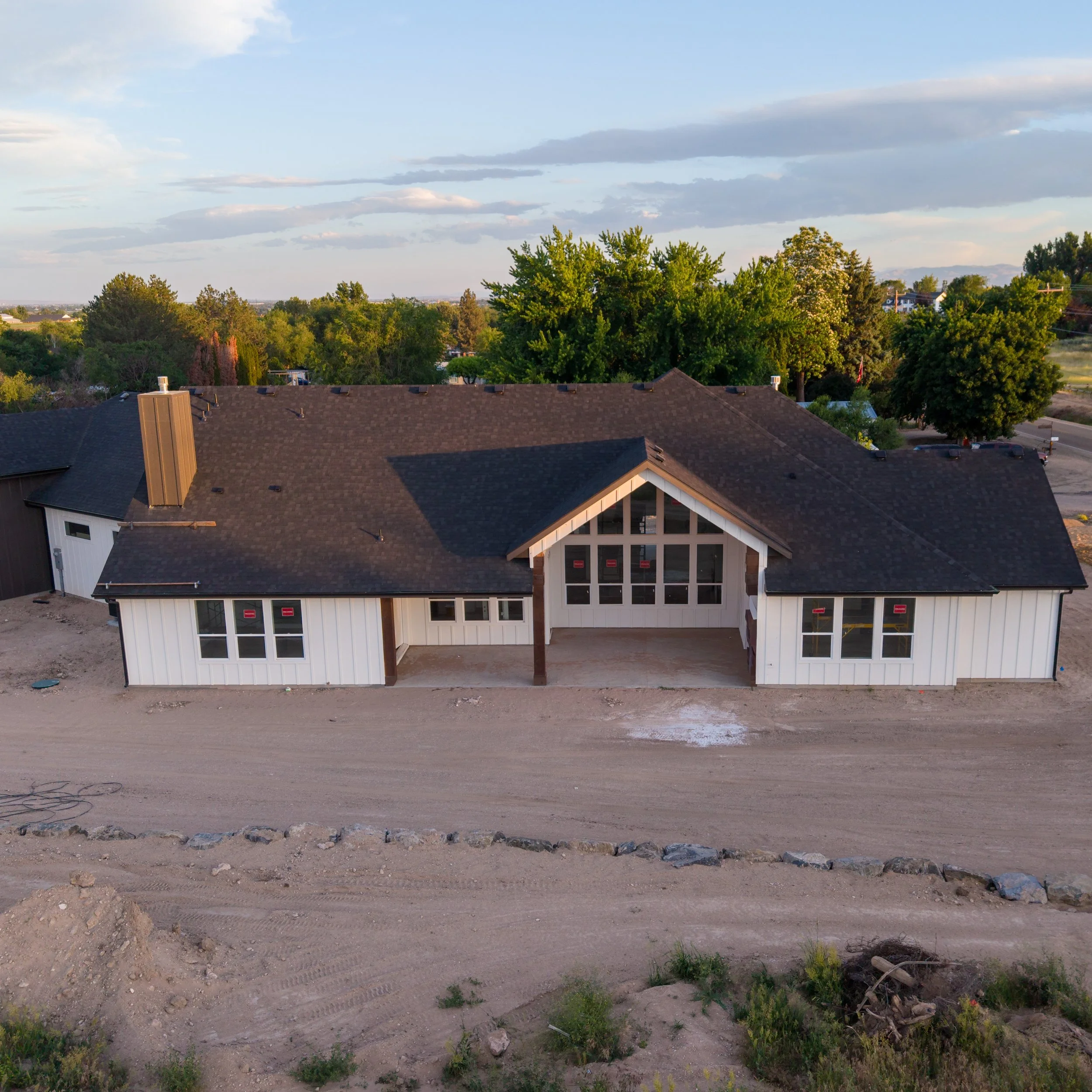 A new house under construction with a partially finished exterior, surrounded by a dirt lot and some green vegetation, with trees and houses in the background under a partly cloudy sky.