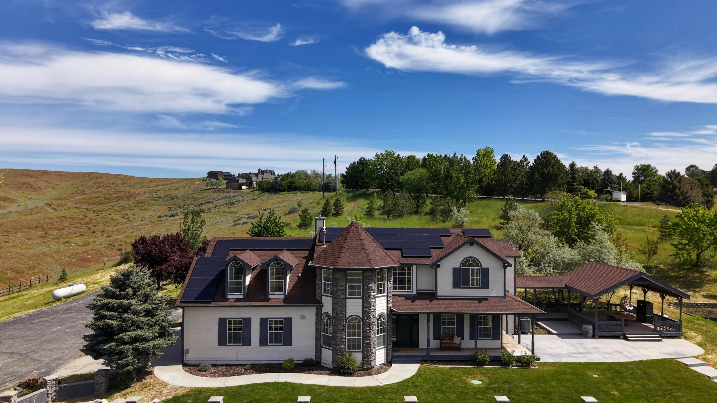 A large house with solar panels on the roof nestled in a hilly, semi-rural landscape under a partly cloudy sky, with greenery and trees surrounding the house.