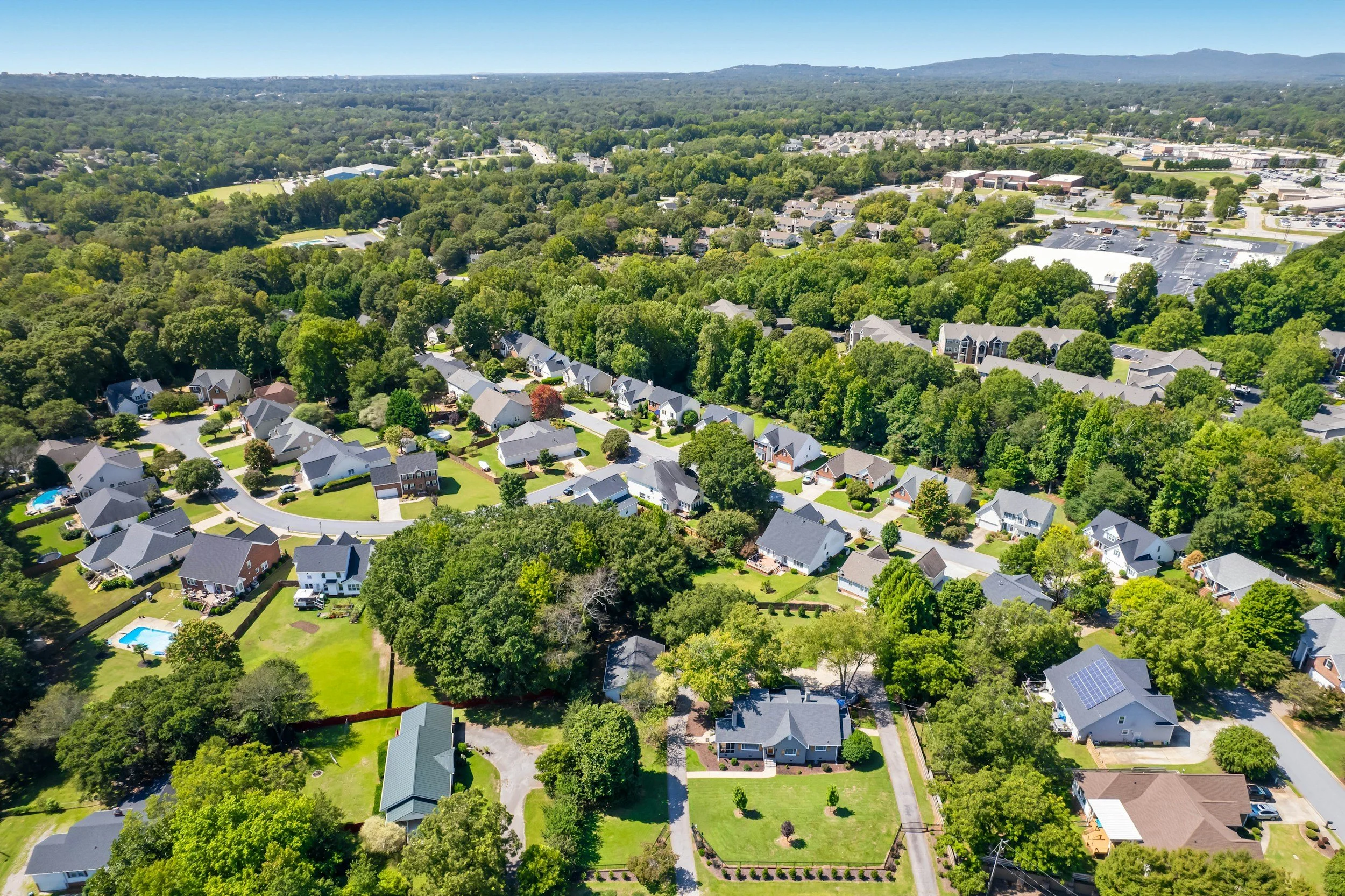 Aerial view of a residential neighborhood with houses surrounded by trees and greenery, some with swimming pools, and a commercial area with parking lots in the background, under a clear blue sky.