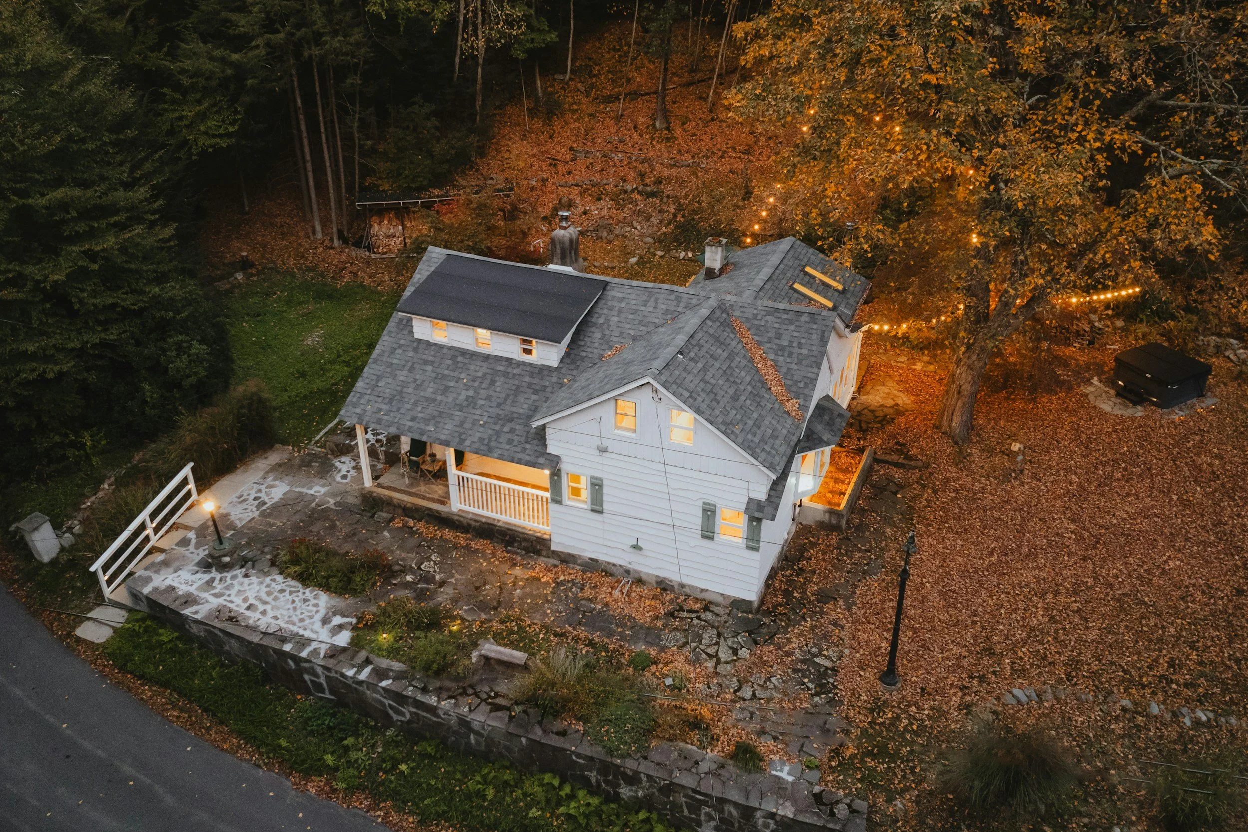 Aerial view of a two-story house with a gray roof surrounded by trees in autumn, with some lights glowing from inside the house.