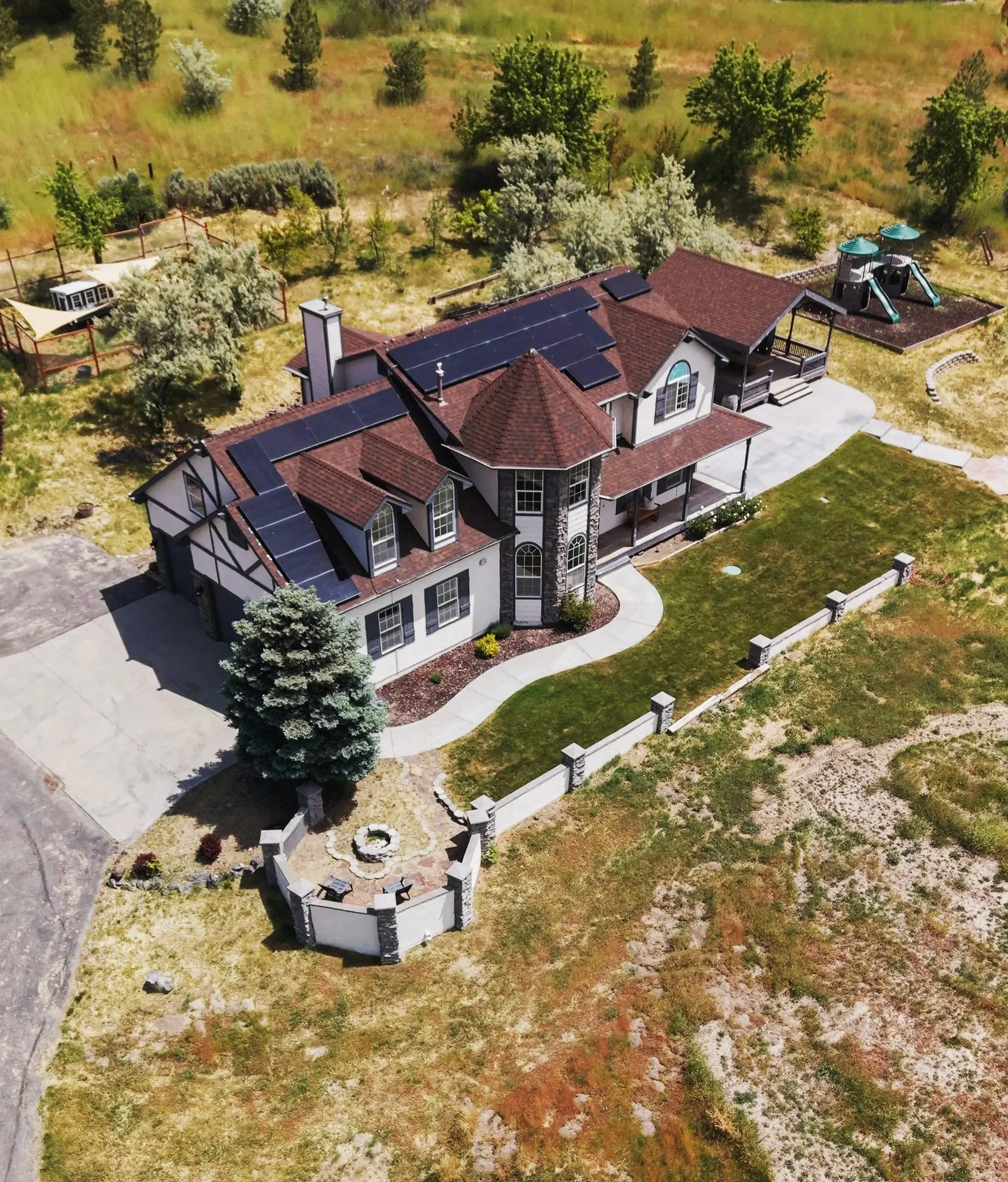 Aerial view of a suburban house with solar panels on the roof, in a grassy backyard with trees and a playground, surrounded by a white fence.