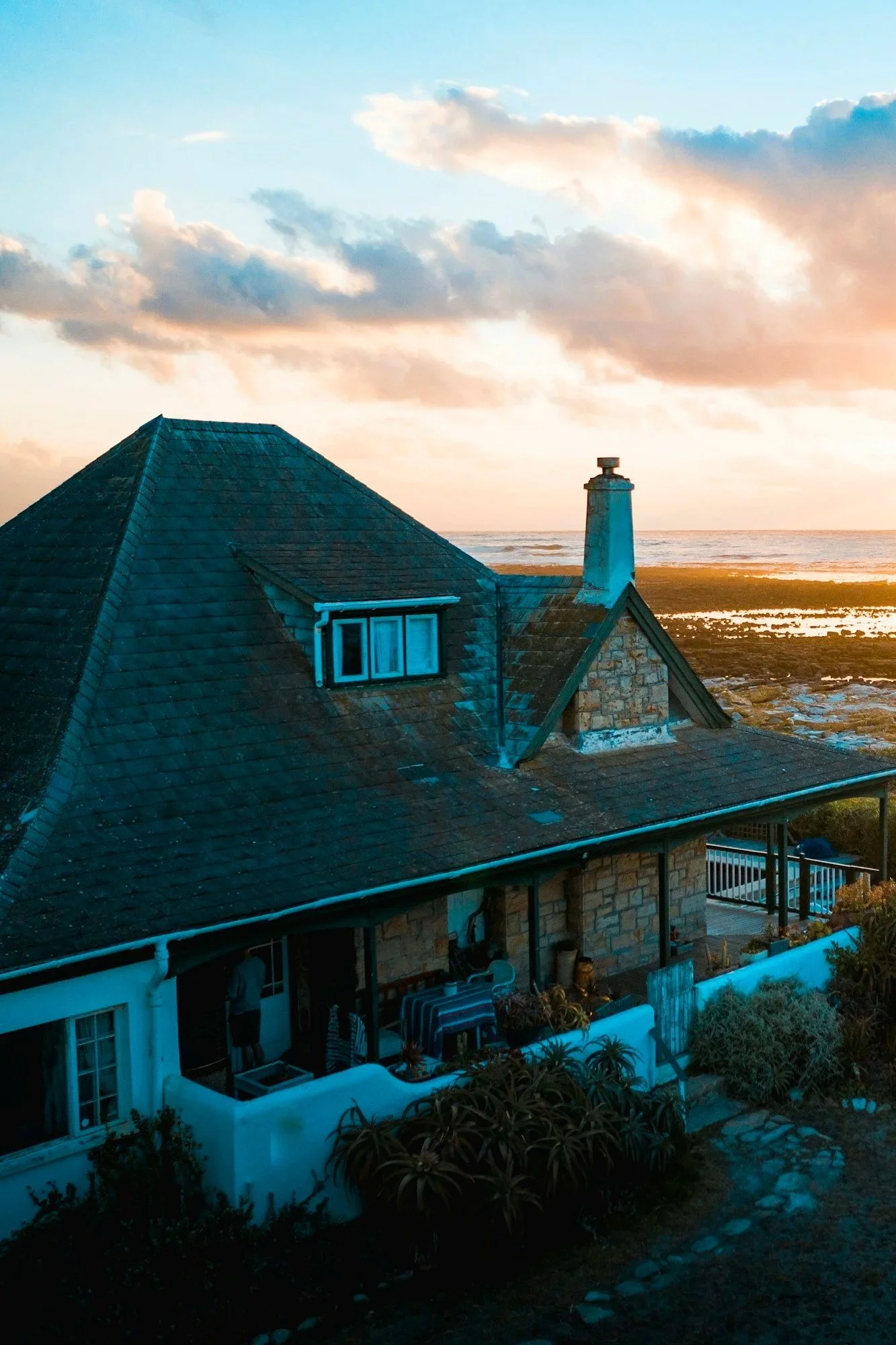 A house with a steep attic roof and a brick chimney by the coast during sunset, with the ocean and cloudy sky in the background.