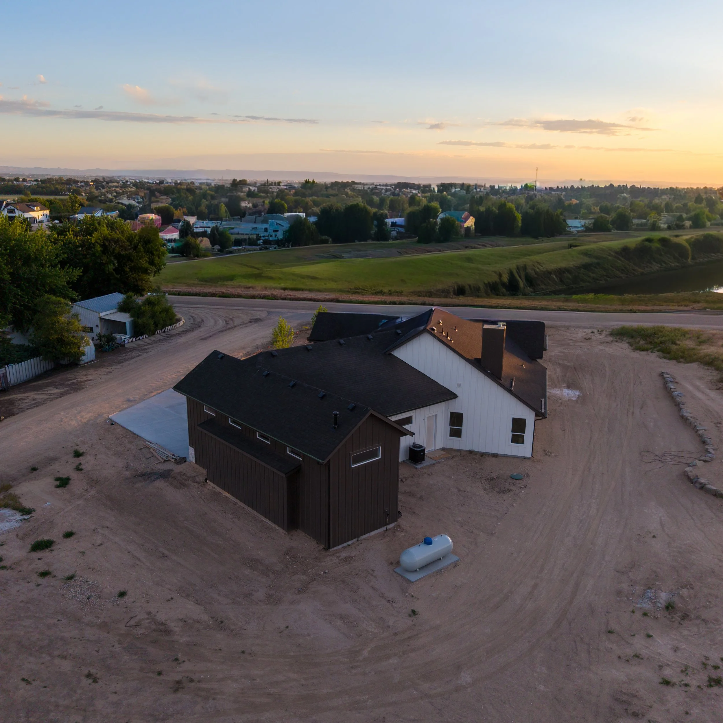 A residential area with a house under construction, surrounded by dirt and grass, with a propane tank in the yard and a distant view of a town under a partly cloudy sky at sunset.