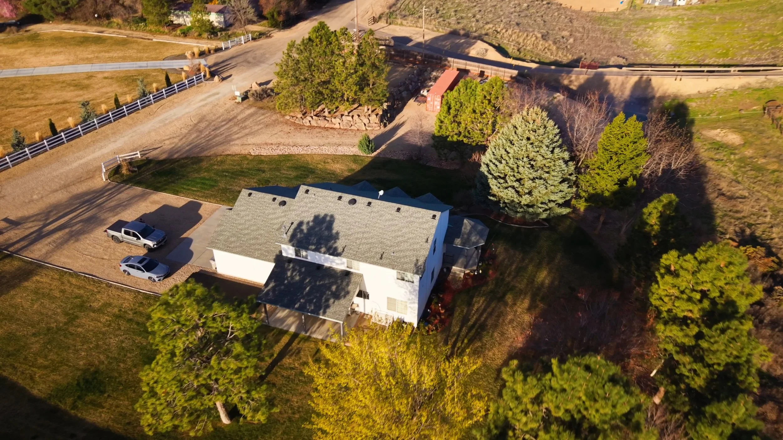 Aerial view of a house with a gray roof, surrounded by trees and a yard with two cars parked in the driveway. There are open grassy areas and a road with a white fence nearby.