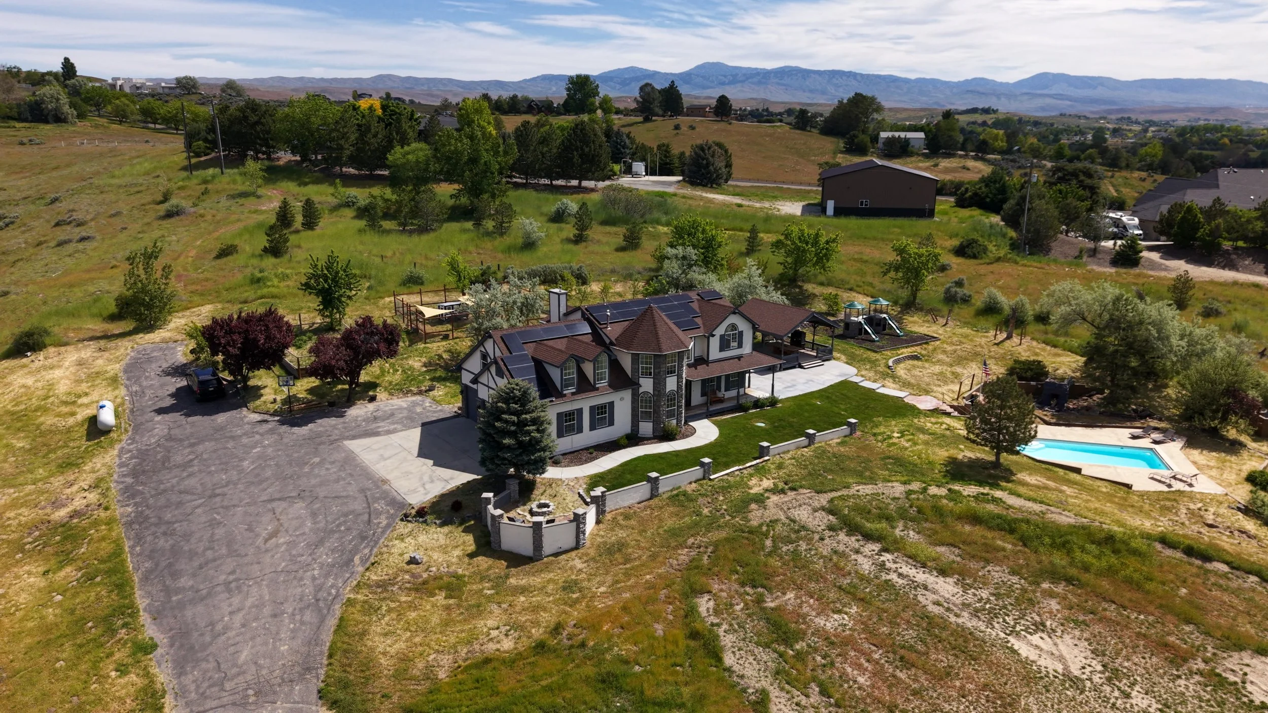 An aerial view of a large two-story house with solar panels on the roof, a circular driveway, a backyard with a swimming pool, and surrounding greenery and trees. In the background, there are hills and open fields under a partly cloudy sky.