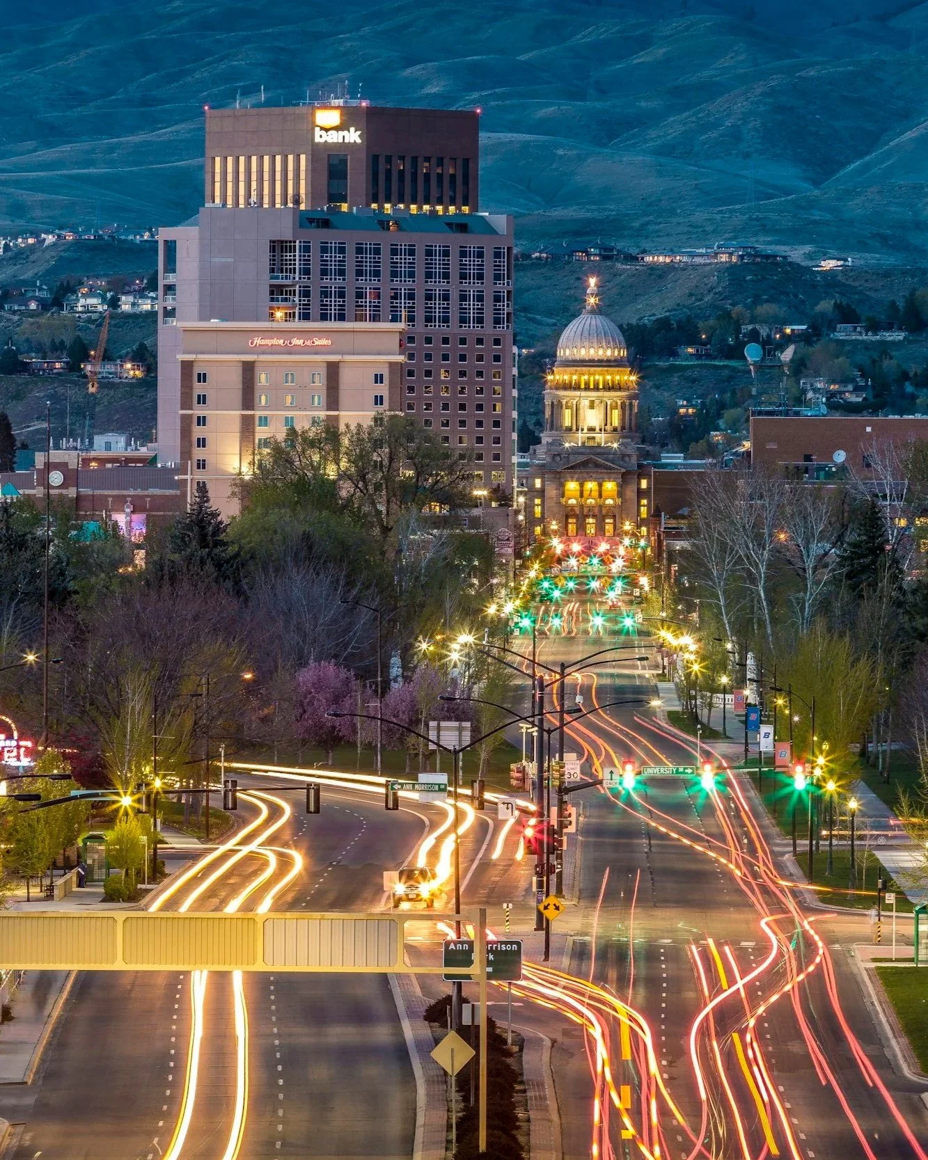Nighttime cityscape with a busy street with moving vehicle light trails, modern buildings including a bank and a government building with a dome, hills in the background, and illuminated signs