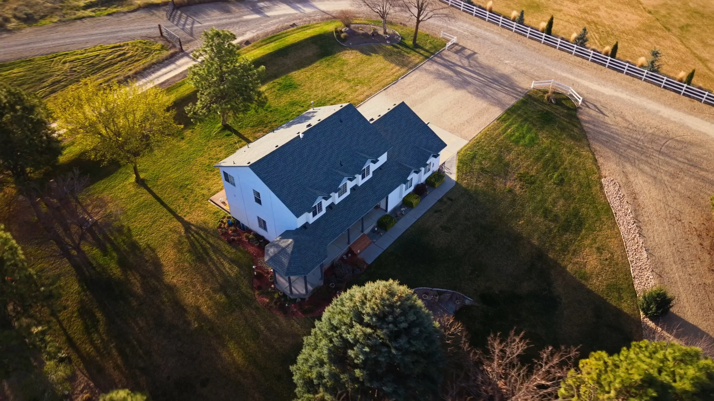 An aerial view of a house with a gray roof, surrounded by a well-maintained lawn, trees, and a dirt driveway, with fencing nearby.