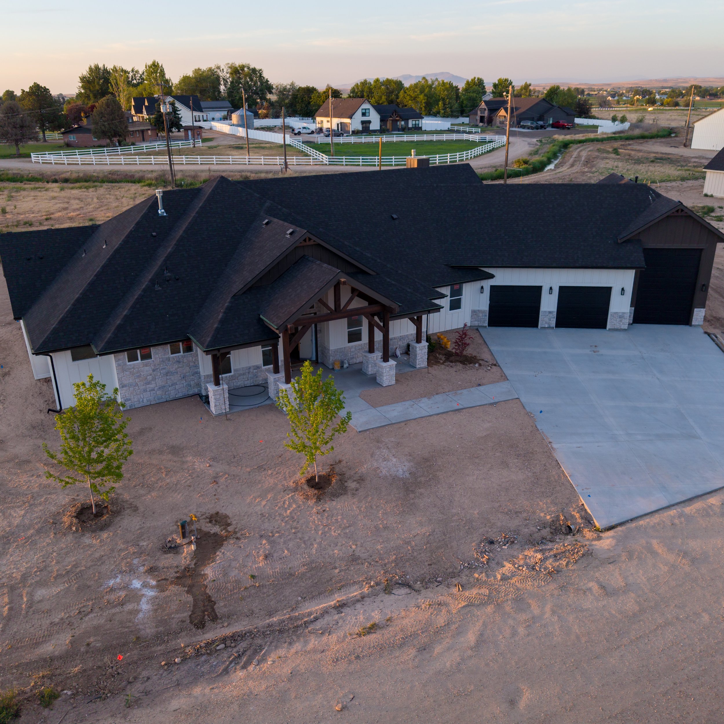 A residential area with new construction homes, small trees, and a dirt yard under development, with mountains in the background during evening light.