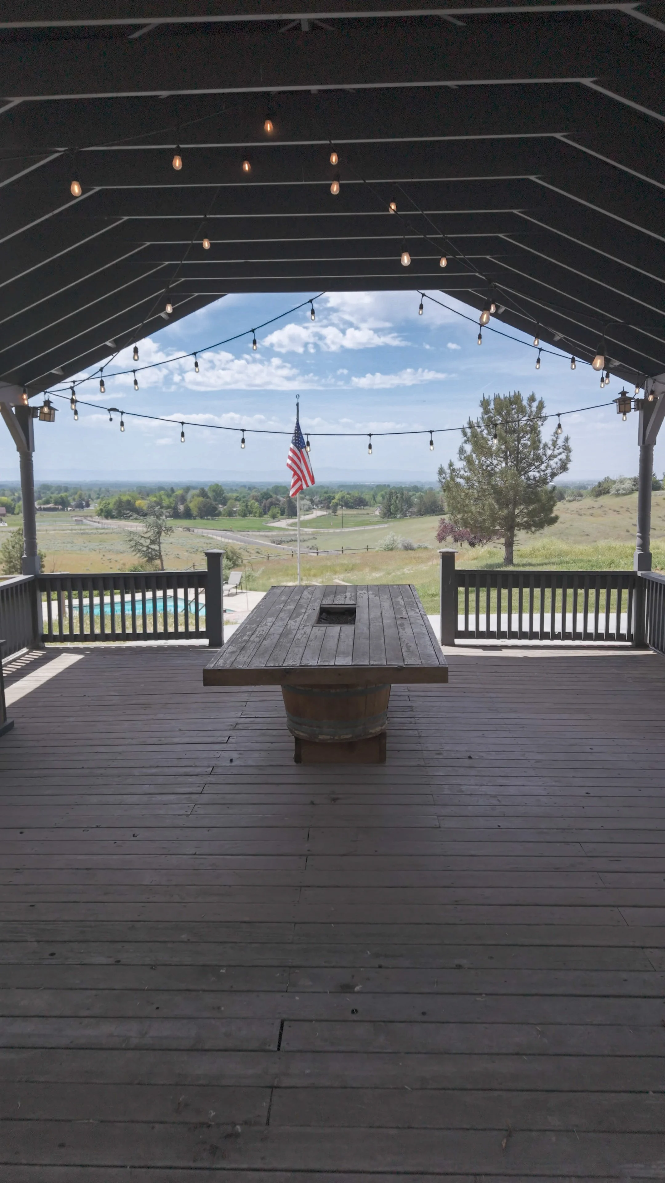 A view from a covered porch with string lights, an American flag, a wooden table, and a deck overlooking a rural landscape with trees, fields, and a swimming pool.