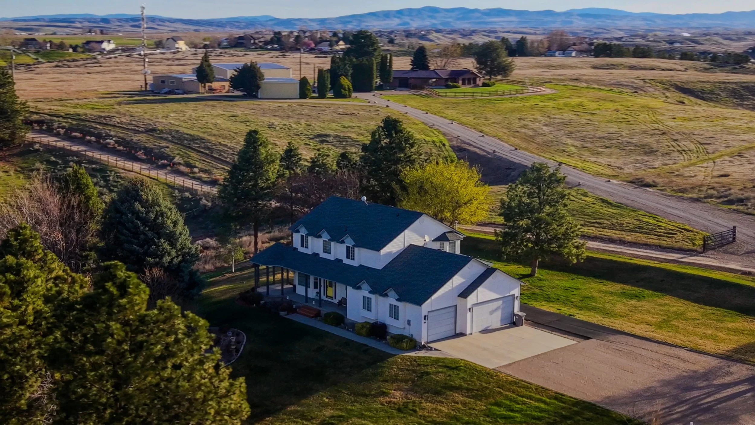 A white two-story house with a sloped roof and attached garage in a rural area with open fields, trees, and distant mountains.