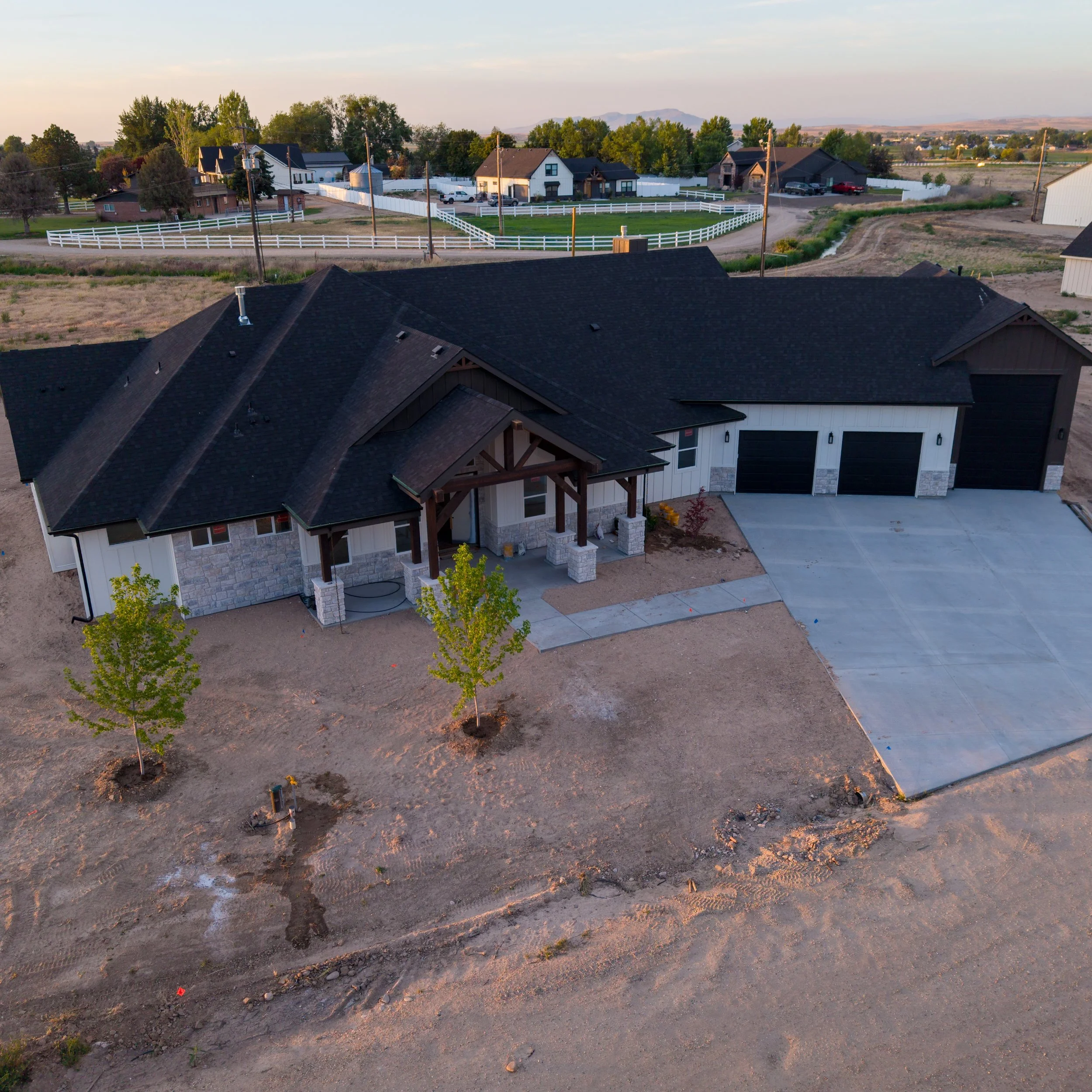 A partially constructed house with a dark roof, stone and siding exterior, and a concrete driveway, with trees and other houses in the background.