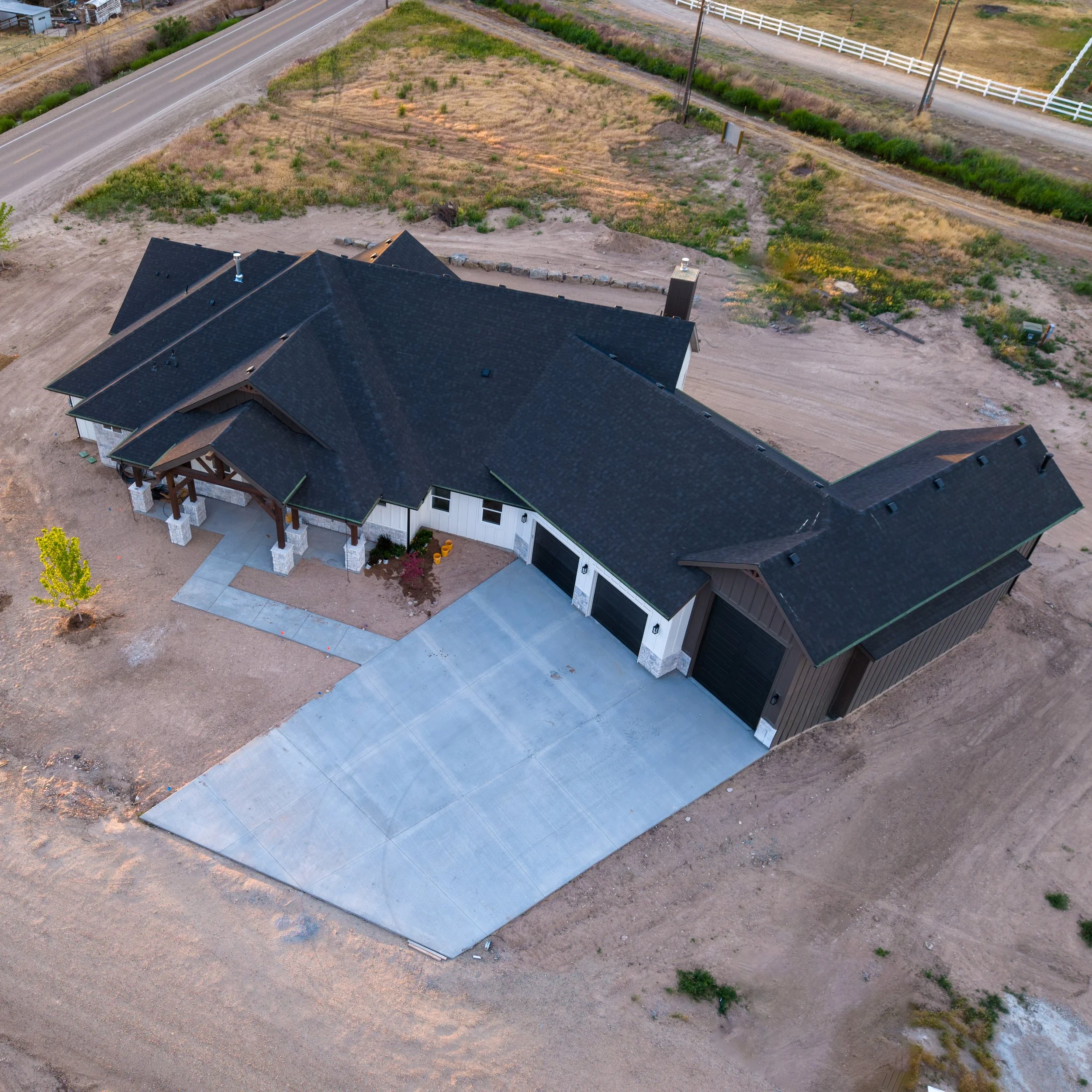 Aerial view of a house with a new concrete driveway, surrounded by dirt and grass patches, with a road and fenced area in the background.