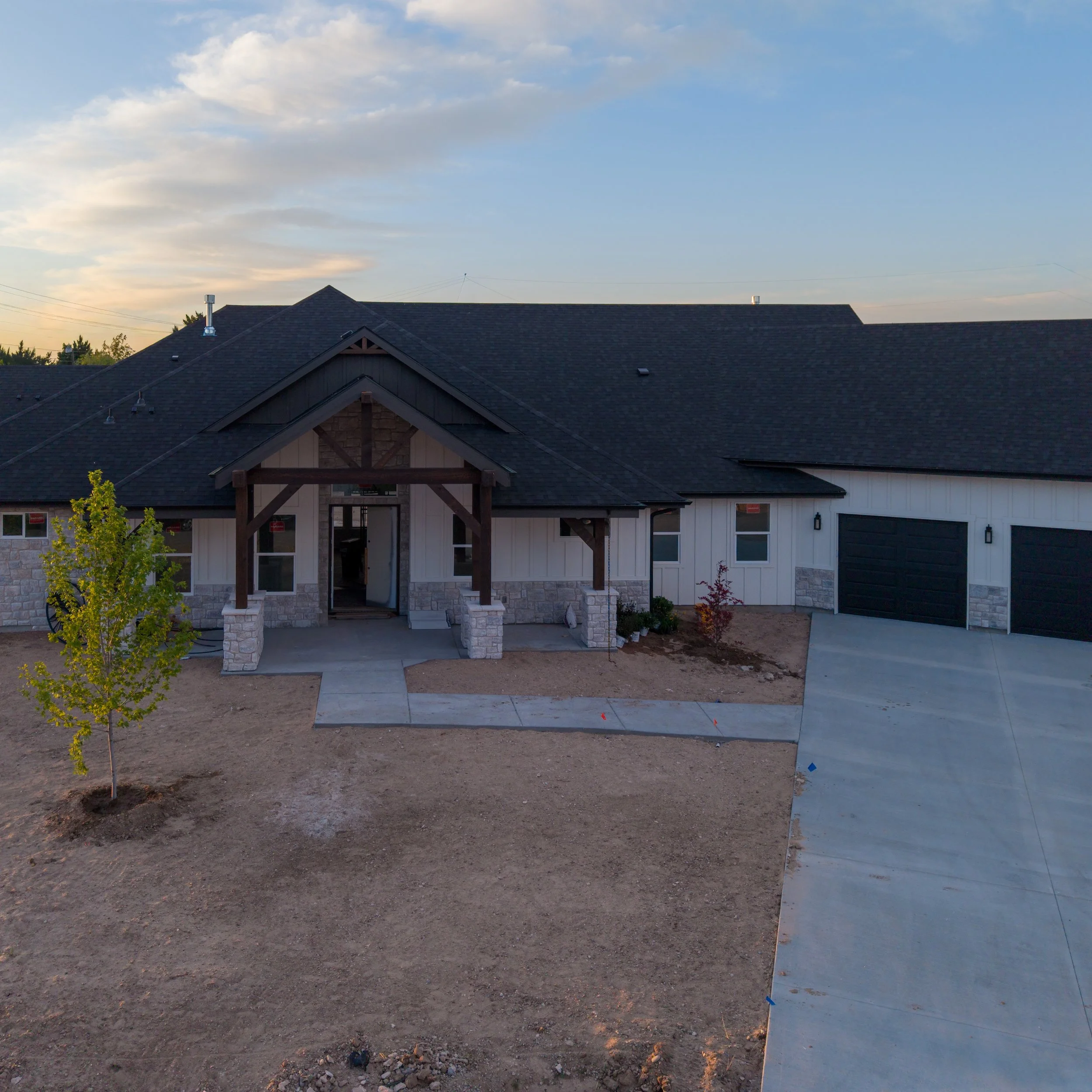 Newly constructed house with a black shingled roof, front porch, stone accents, and adjacent driveway and landscaping in early evening light.