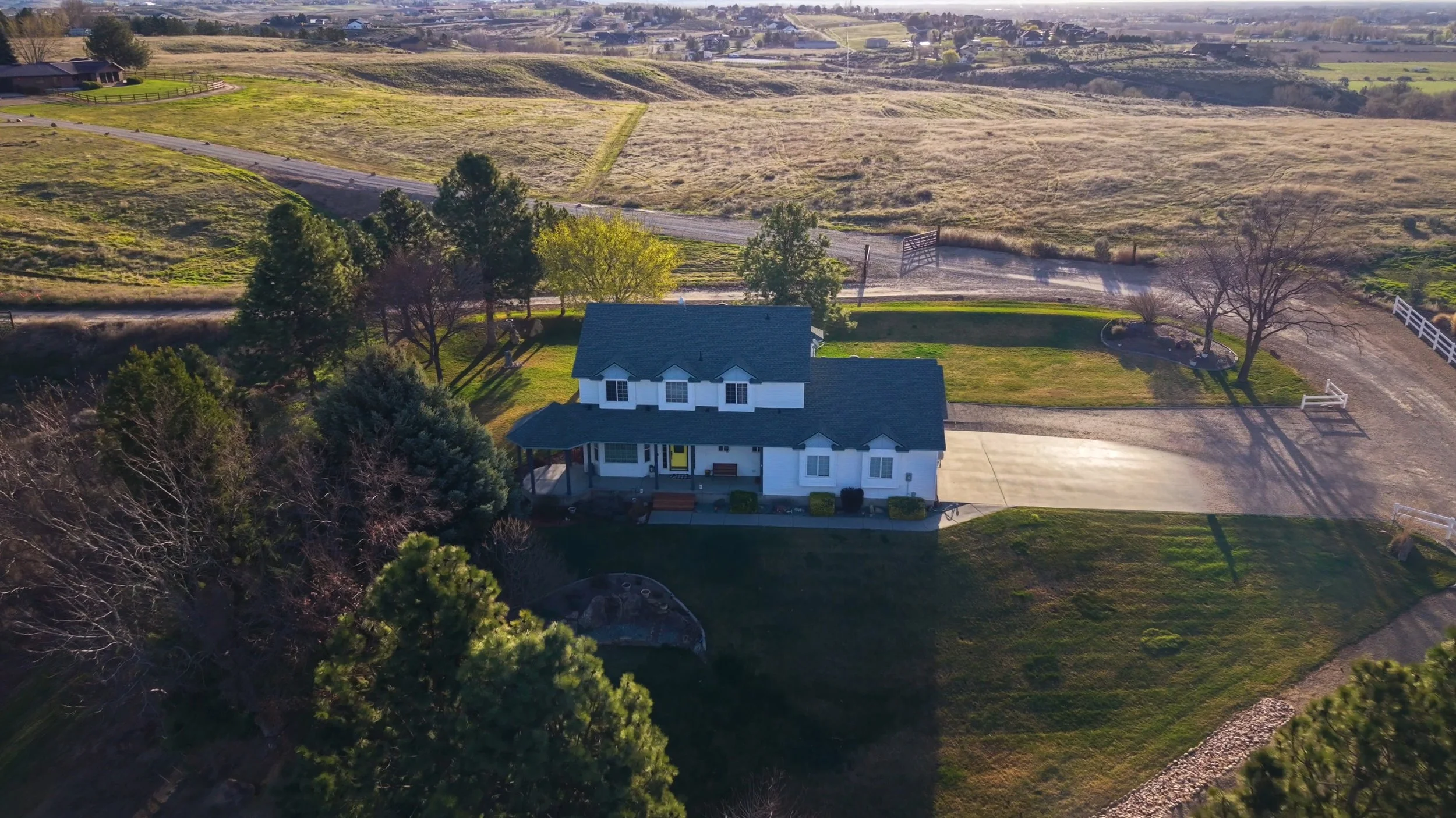 An aerial view of a white two-story house with a dark roof, surrounded by trees and a well-maintained lawn, with farmland and open fields in the background.