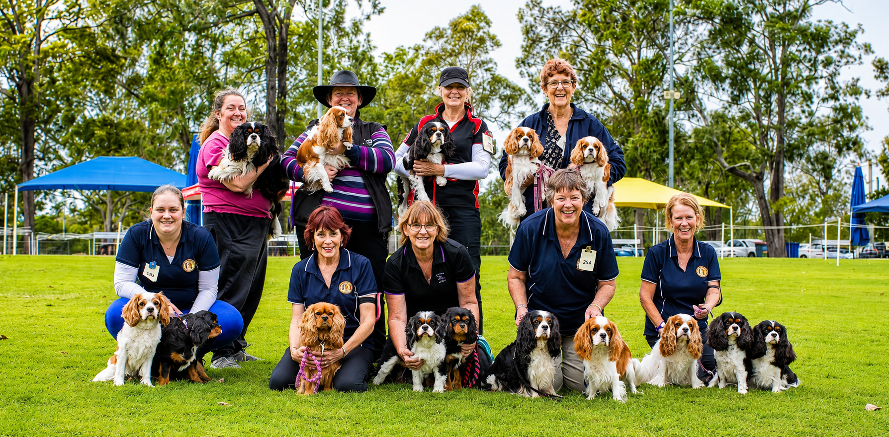 A group of women with their dogs on grass in a park, smiling for a photo, with tents and trees in the background.