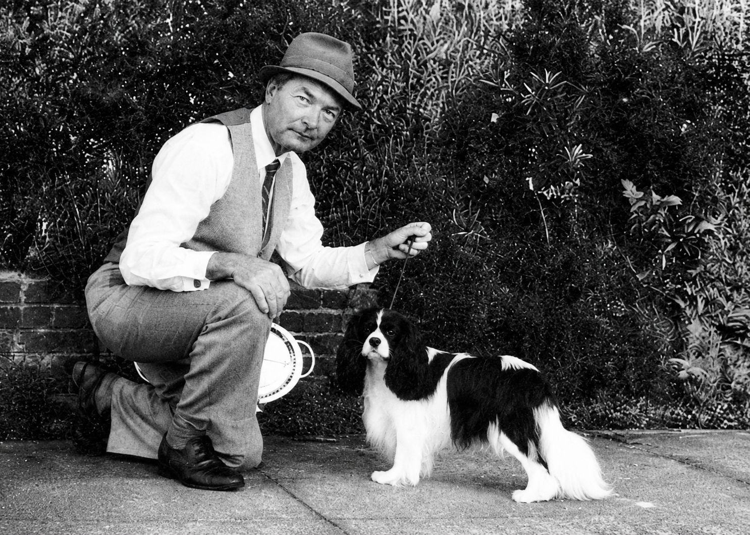 A man wearing a hat, white shirt, vest, and tie squats next to a black and white dog on a leash. They are outdoors, in front of a bushy background, on a sidewalk.