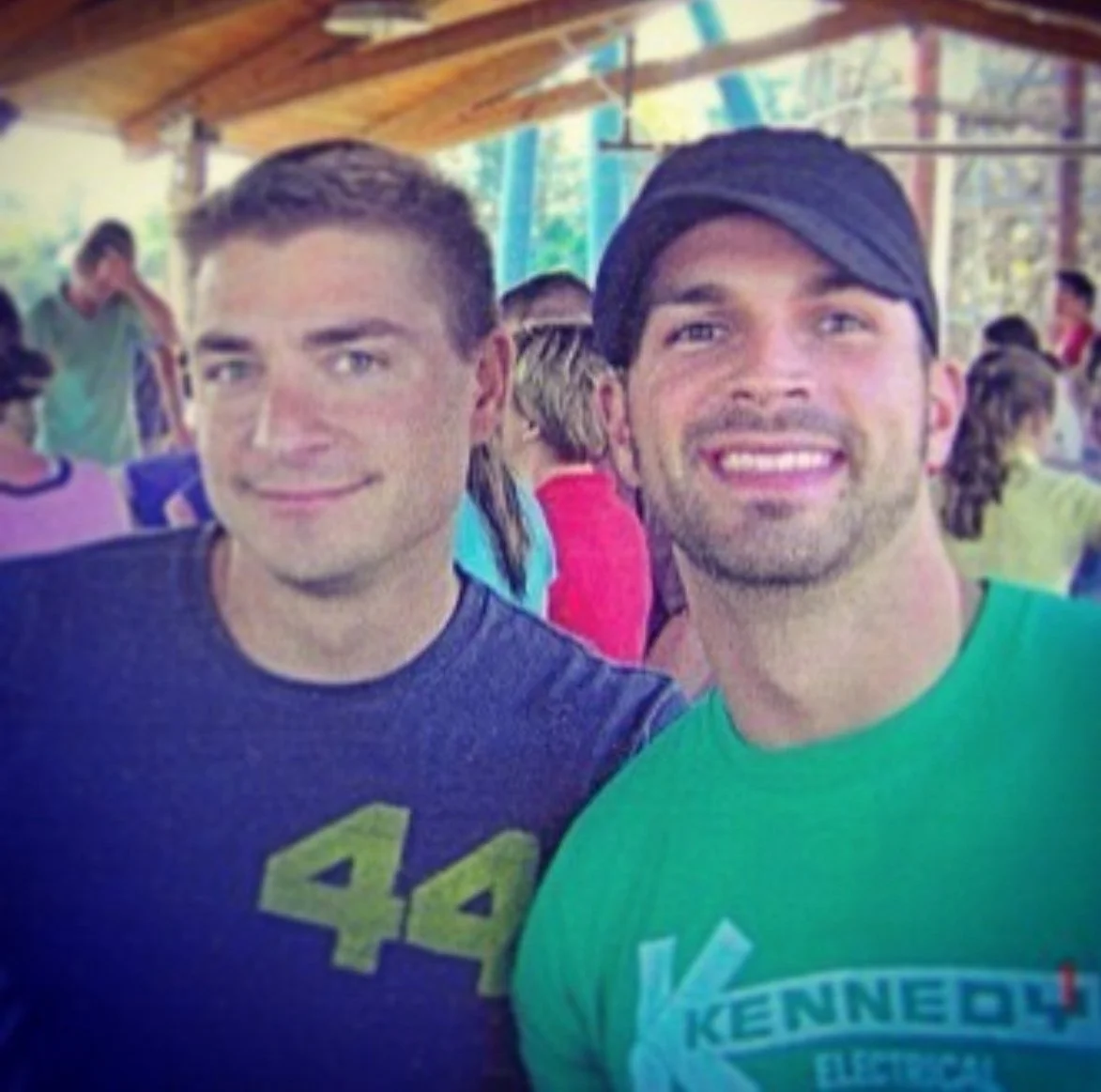 Jeremy Sueksdorf, wearing a navy shirt with a yellow '44' on the left and Matthew Fior wearing a black cap and green shirt on the right posing for a photo at a social theme park with other people in the background.