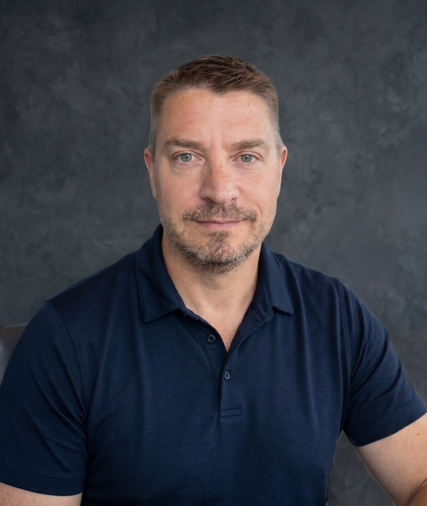 Jeremy Sueksdorf with a short light brown hair and stubble, wearing a navy blue polo shirt, sitting against a dark textured background, looking at the camera with smile.