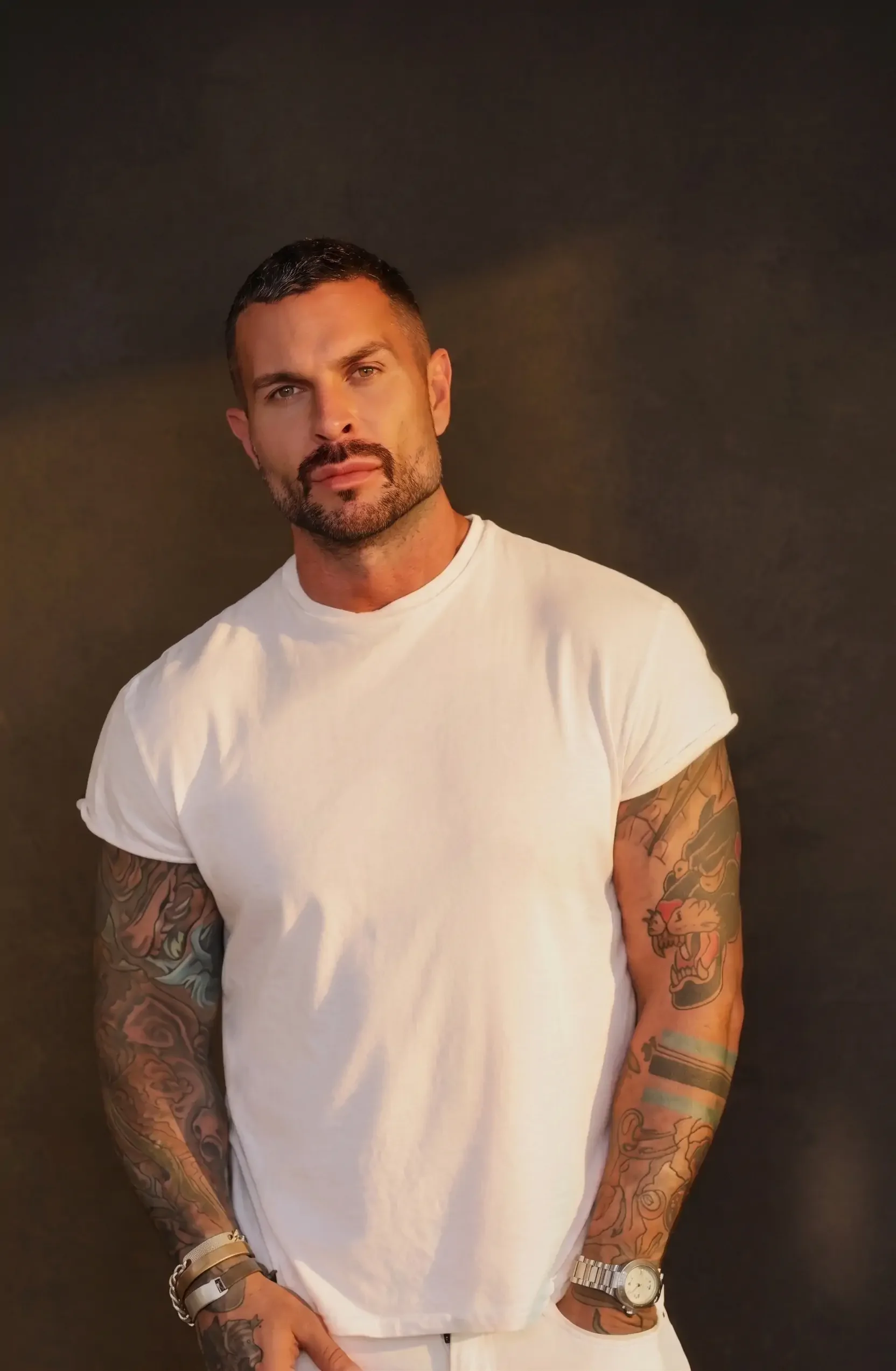 MGF & Partners Founder, Matthew G. Fior with short dark hair, a beard and tattoos on both arms wearing a white t-shirt, beige pants, and a watch, standing against a dark background.