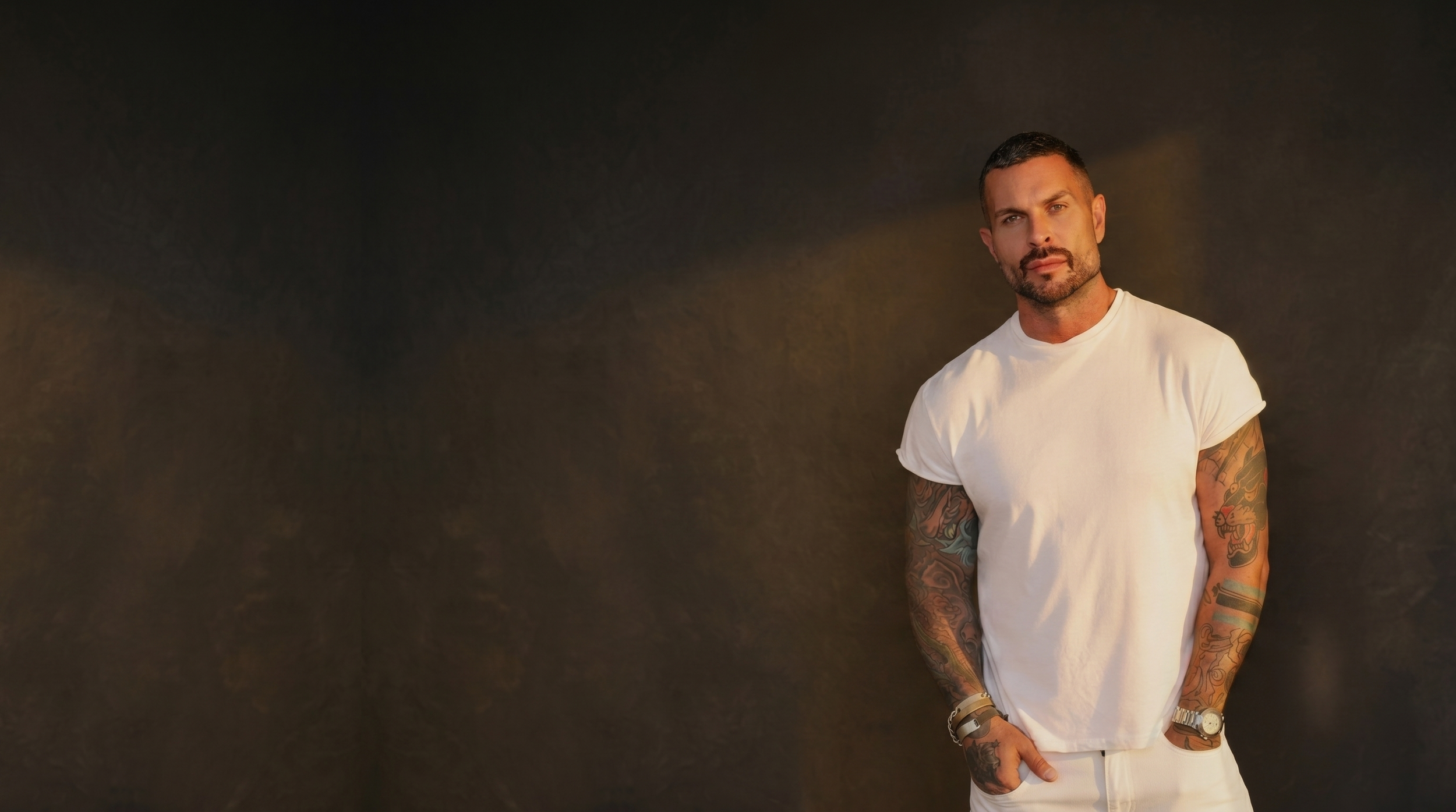 A man with tattoos on his arms is standing against a dark background, wearing a white t-shirt and looking at the camera.