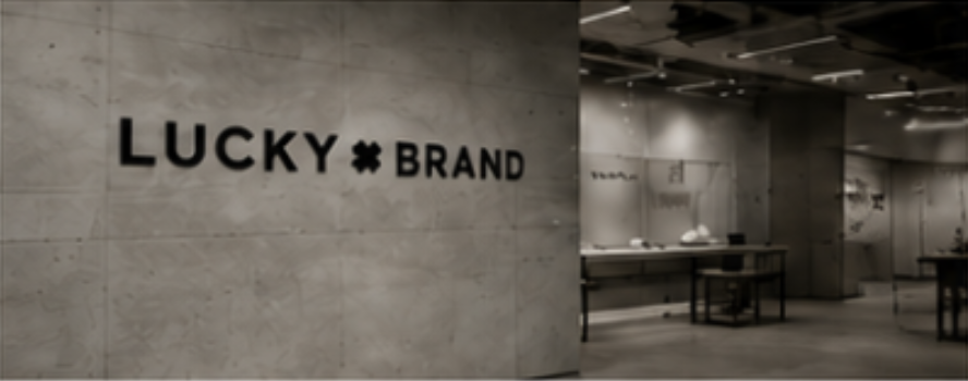 Interior of a retail store with a concrete wall displaying the 'Lucky Brand' logo, and several display tables with merchandise.
