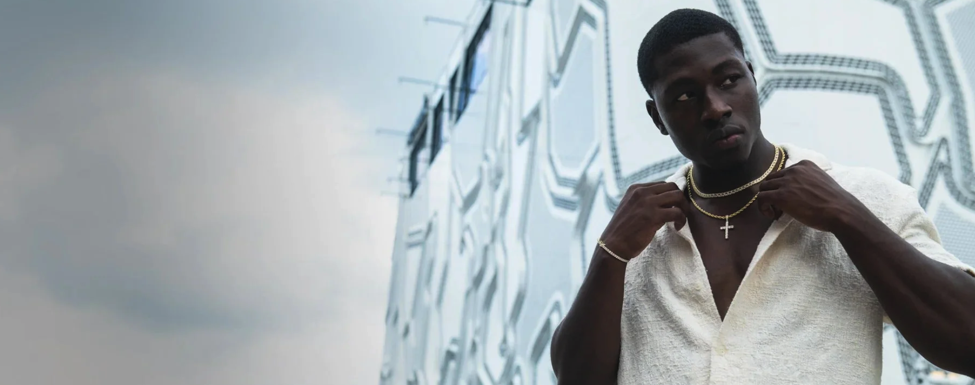 A young man with dark skin and short hair adjusting gold chains around his neck. He is wearing a cream-colored shirt and standing in front of a modern, geometric patterned wall.