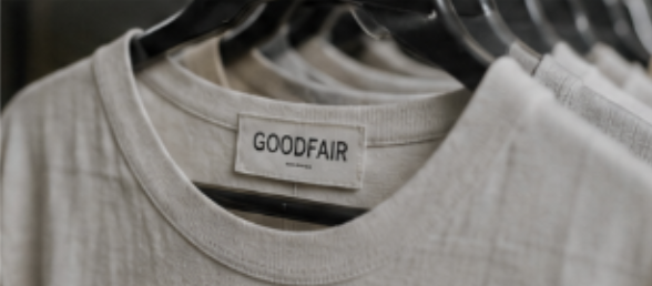 Close-up of a beige t-shirt with a 'GOODFAIR' label, hanging among similar shirts on a rack.