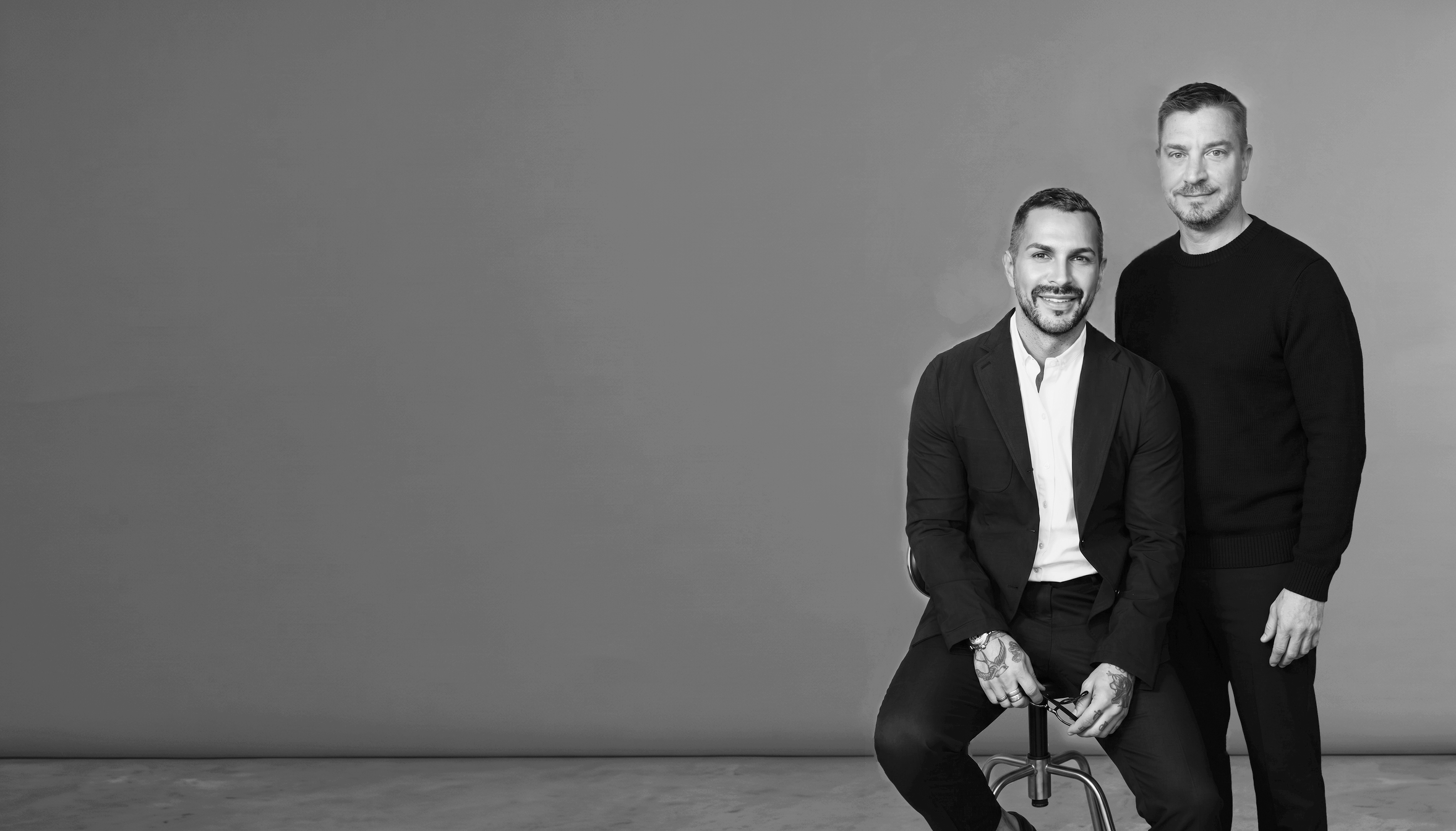 Matthew Fior is seated on a stool, wearing a suit and smiling. Jeremy Suksdorf standing beside him, wearing a dark sweater and also smiling.