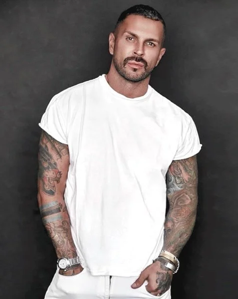 Matthew Gregory Fior wearing a white T-shirt showing his tattoos and jewelry on both wrists, standing against a dark background.