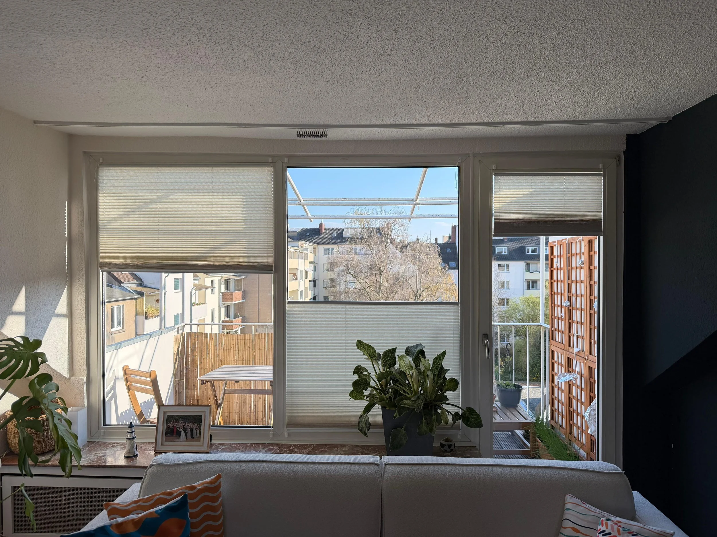 Living room with large window overlooking a balcony and neighboring buildings. Interior decorated with plants, framed photos, and cushions.