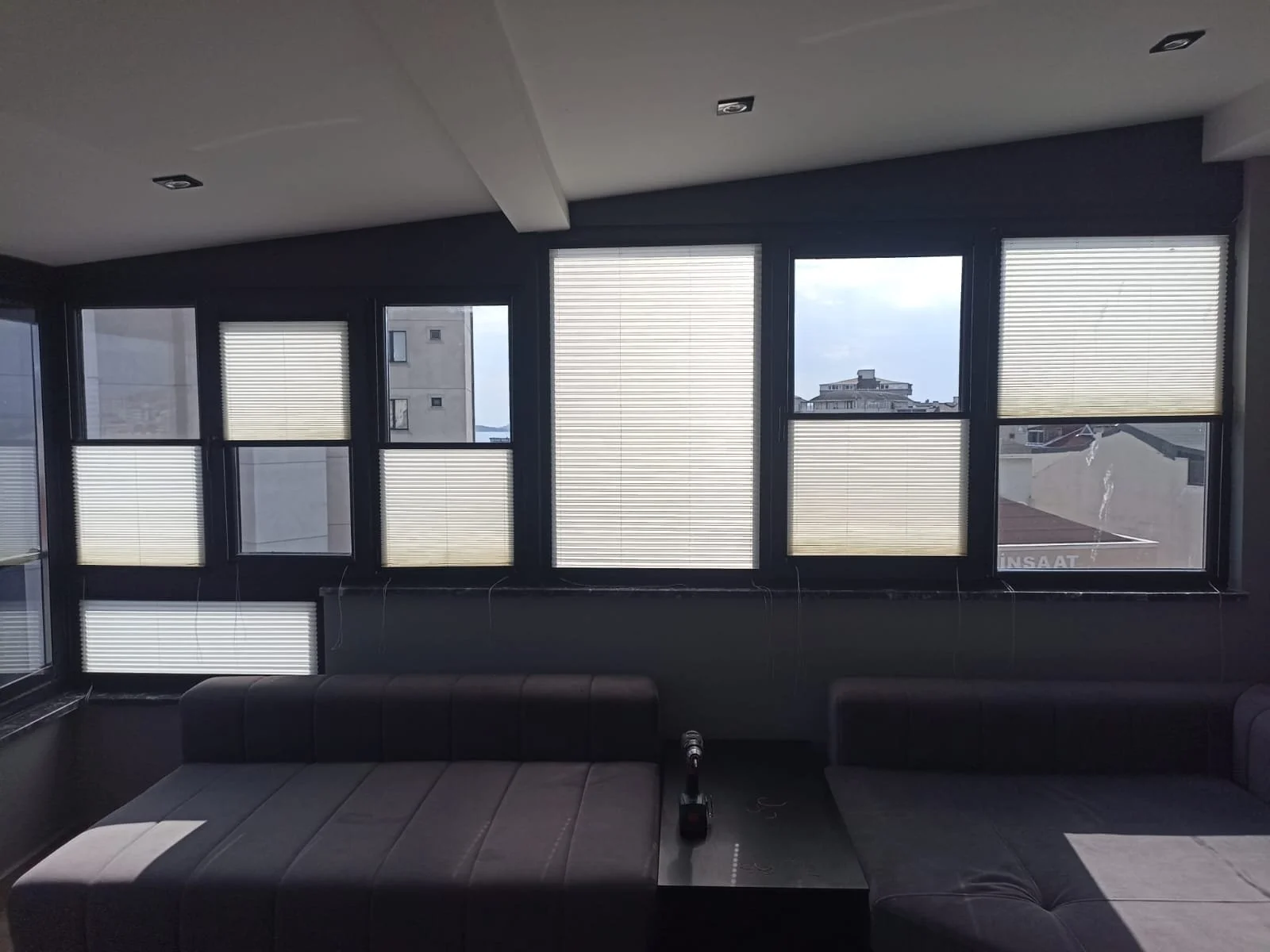 Interior of a modern living room with large windows covered by partially closed blinds, a black sectional sofa, and a view of neighboring buildings outside.