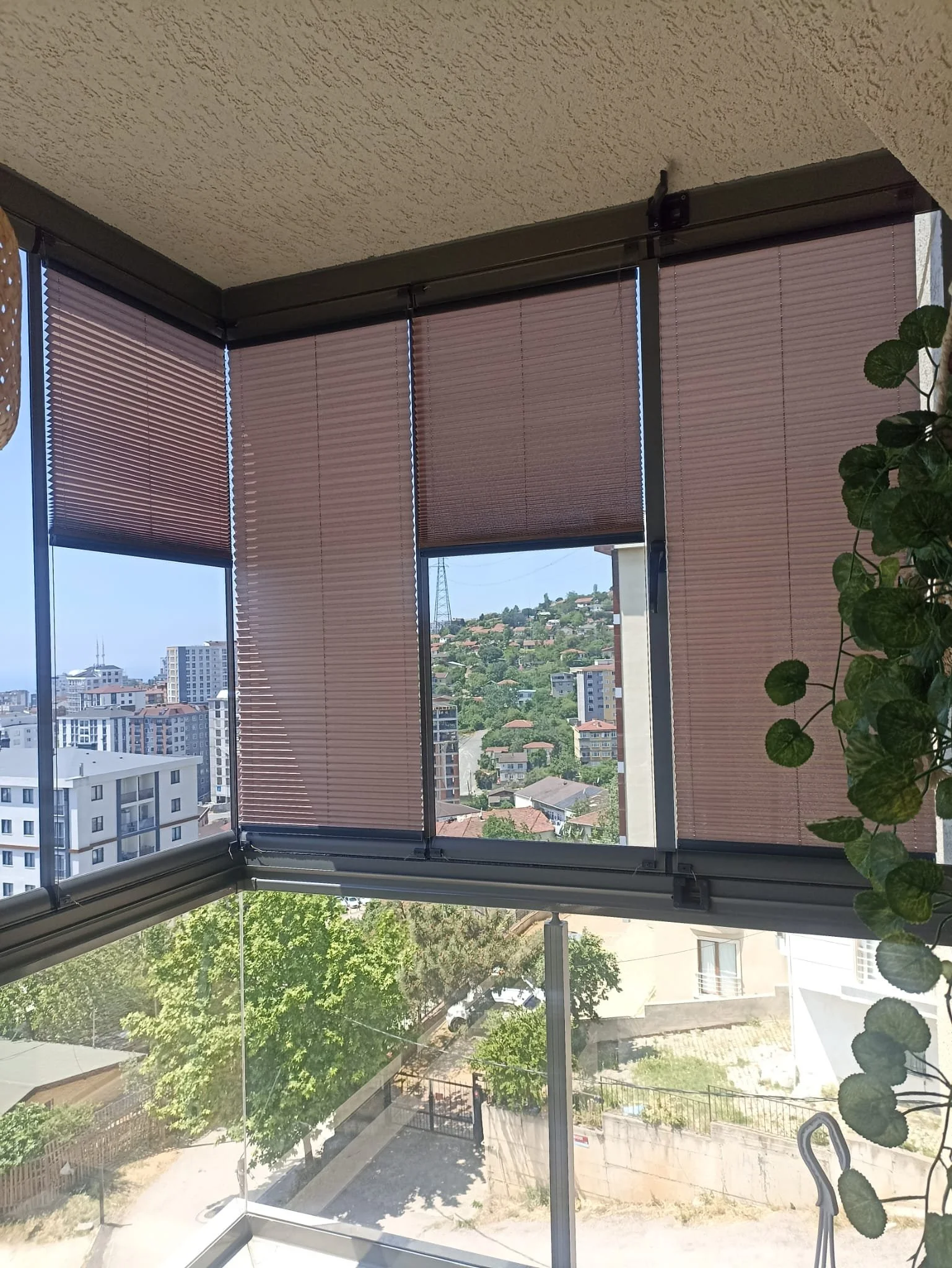 View from a balcony or enclosed room showing cityscape with high-rise buildings, trees, and houses in the distance, with window blinds partially closed.