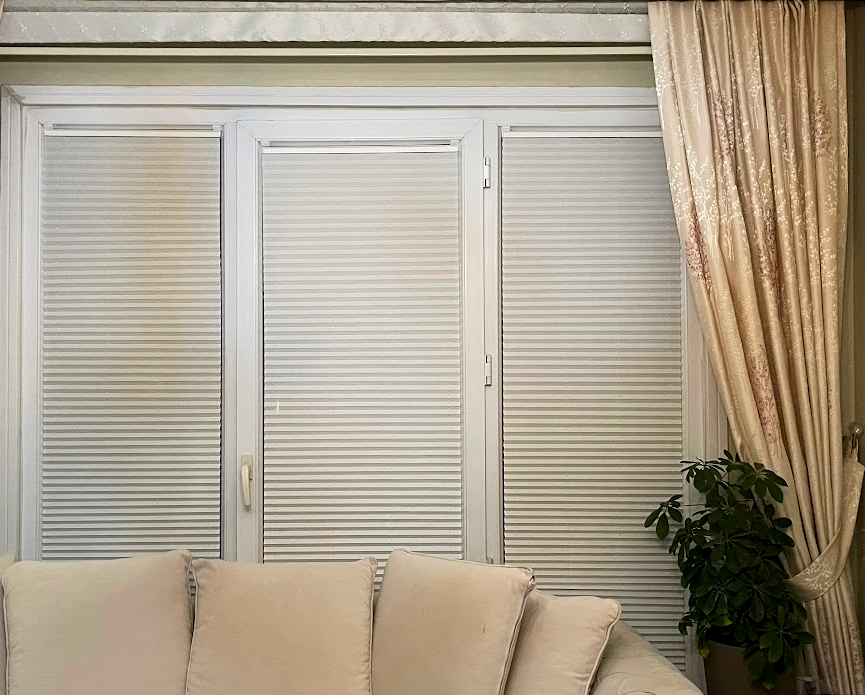 Living room with a white sofa, window with white shutters, and a beige curtain with floral patterns, and a green potted plant on the right side.