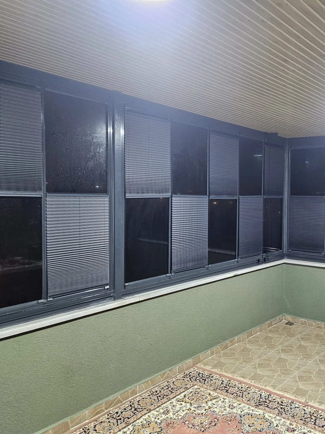 A covered balcony with a green wall, patterned tile flooring, and black-framed windows with partially open horizontal blinds.