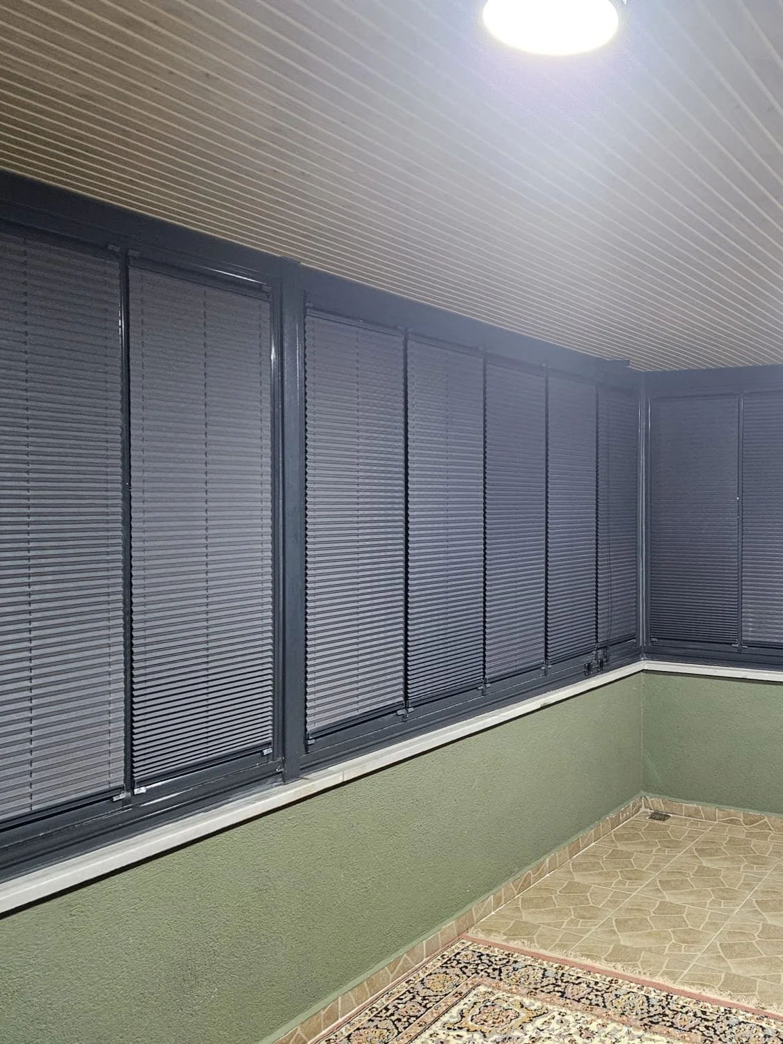 Enclosed balcony with black window shutters, green lower wall, beige tiled floor, ceiling light, and a patterned rug.